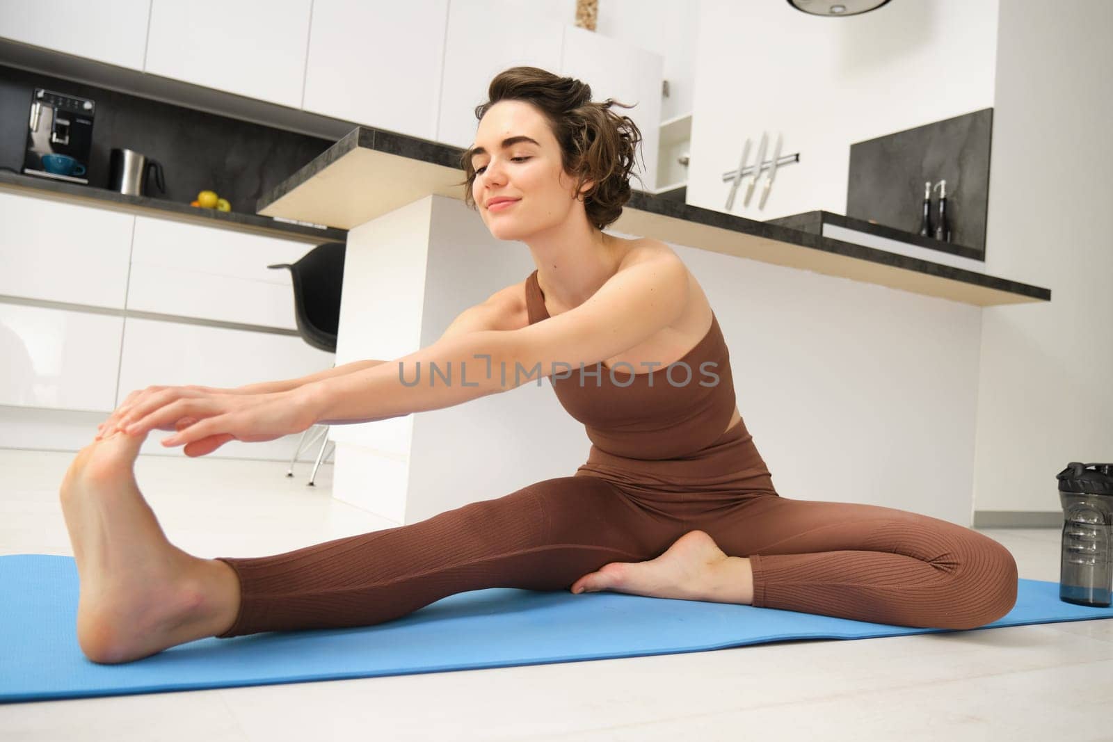 Portrait of brunette girl, fitness instructor, doing warm up training, exercises at home, stretching her legs on yoga mat at home.