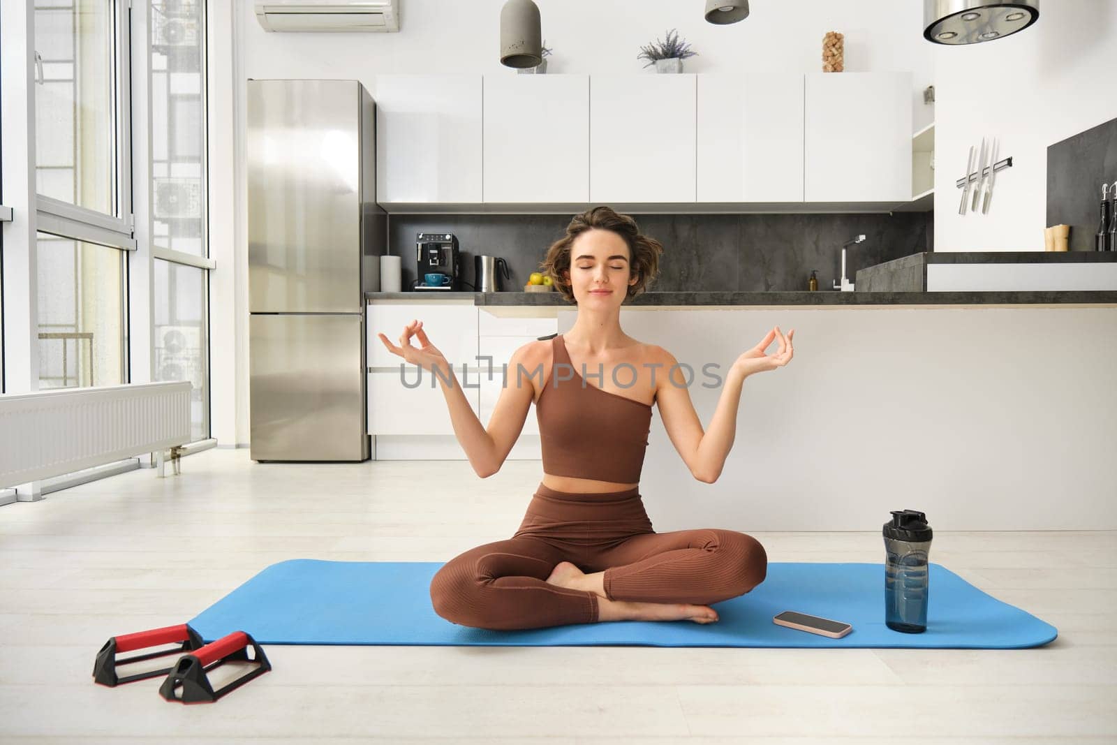 Smiling girl athlete, sportswoman doing yoga at home in activewear, sitting on yoga rubber mat in lotus pose, meditating, practice mindfulness exercises by Benzoix
