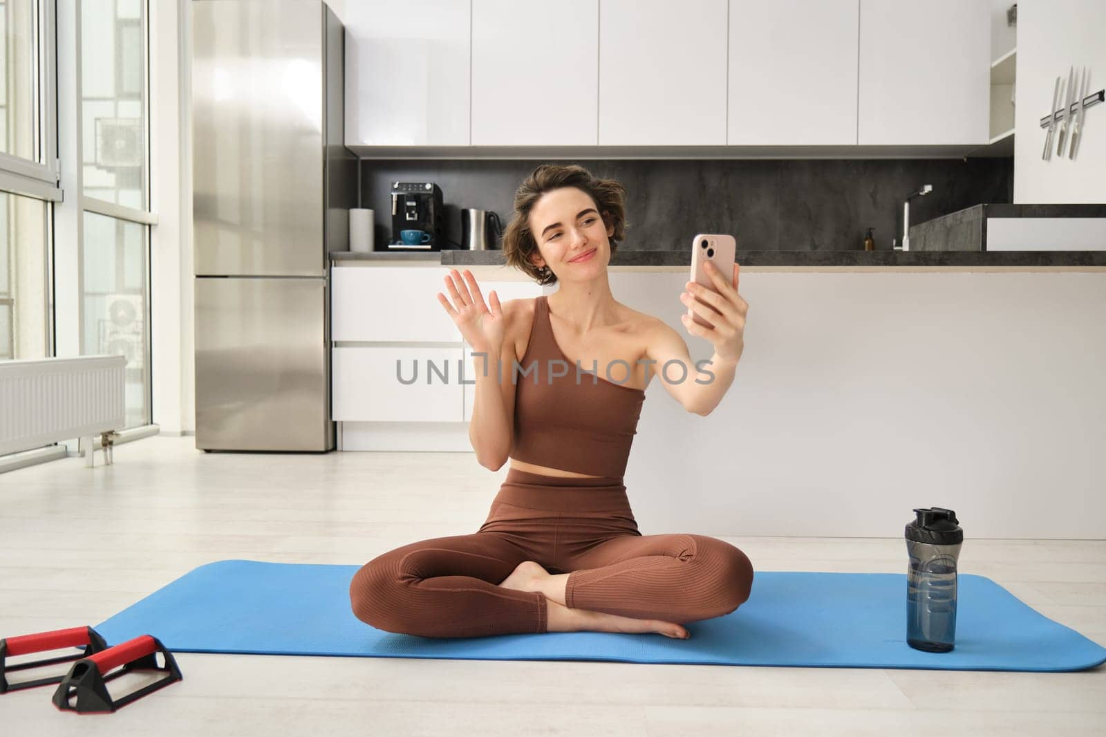 Portrait of young fitness woman, sitting at home on rubber mat, records video on smartphone, doing workout vlog, making live online training session from home by Benzoix