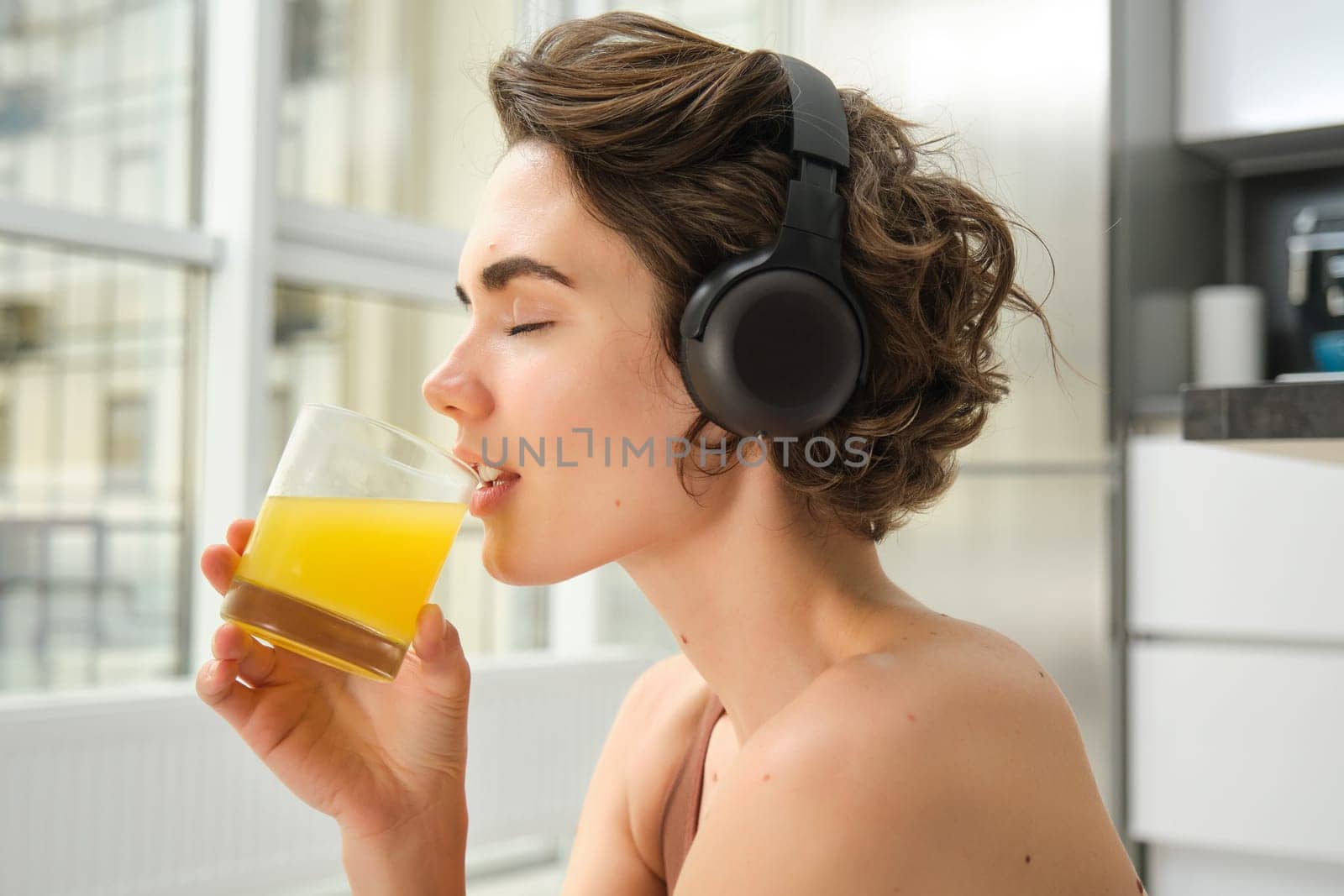 Smiling sportswoman, fitness girl does workout at home, wears headphones, listens music and drinks orange juice after training session.