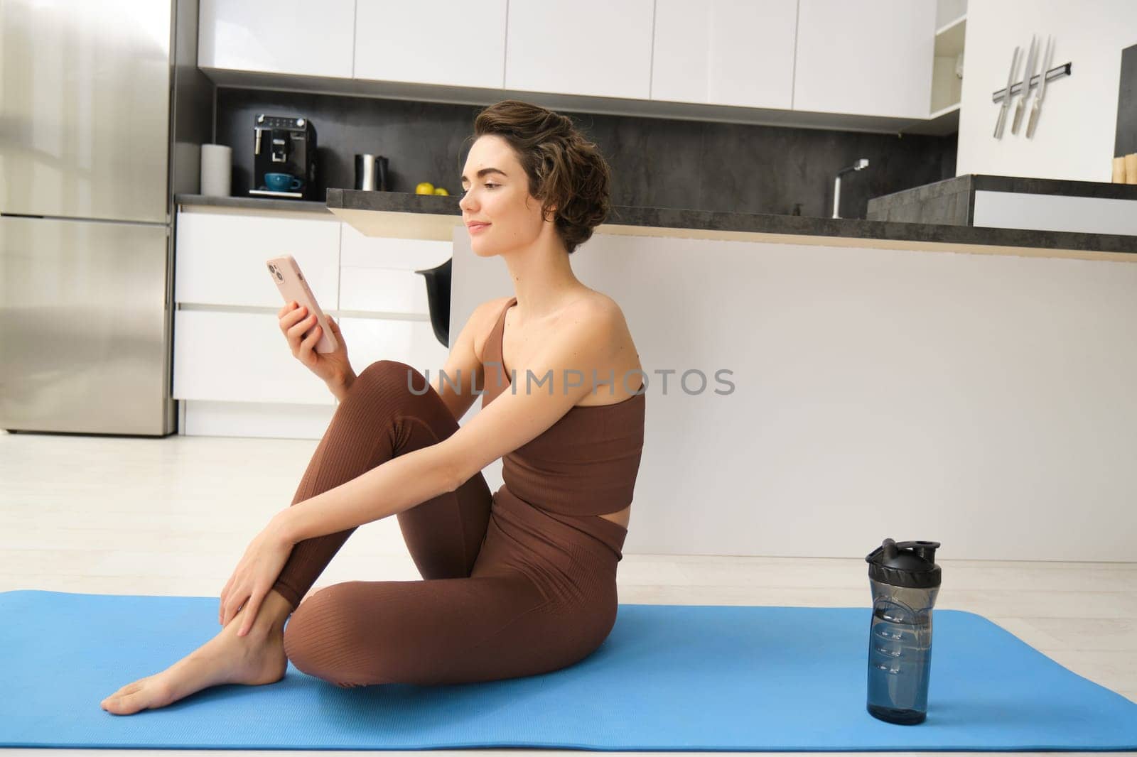 Young fitness instructor looking at her smartphone, sitting on rubber mat at home. Young woman doing workout training, looking at exercise videos on mobile app.