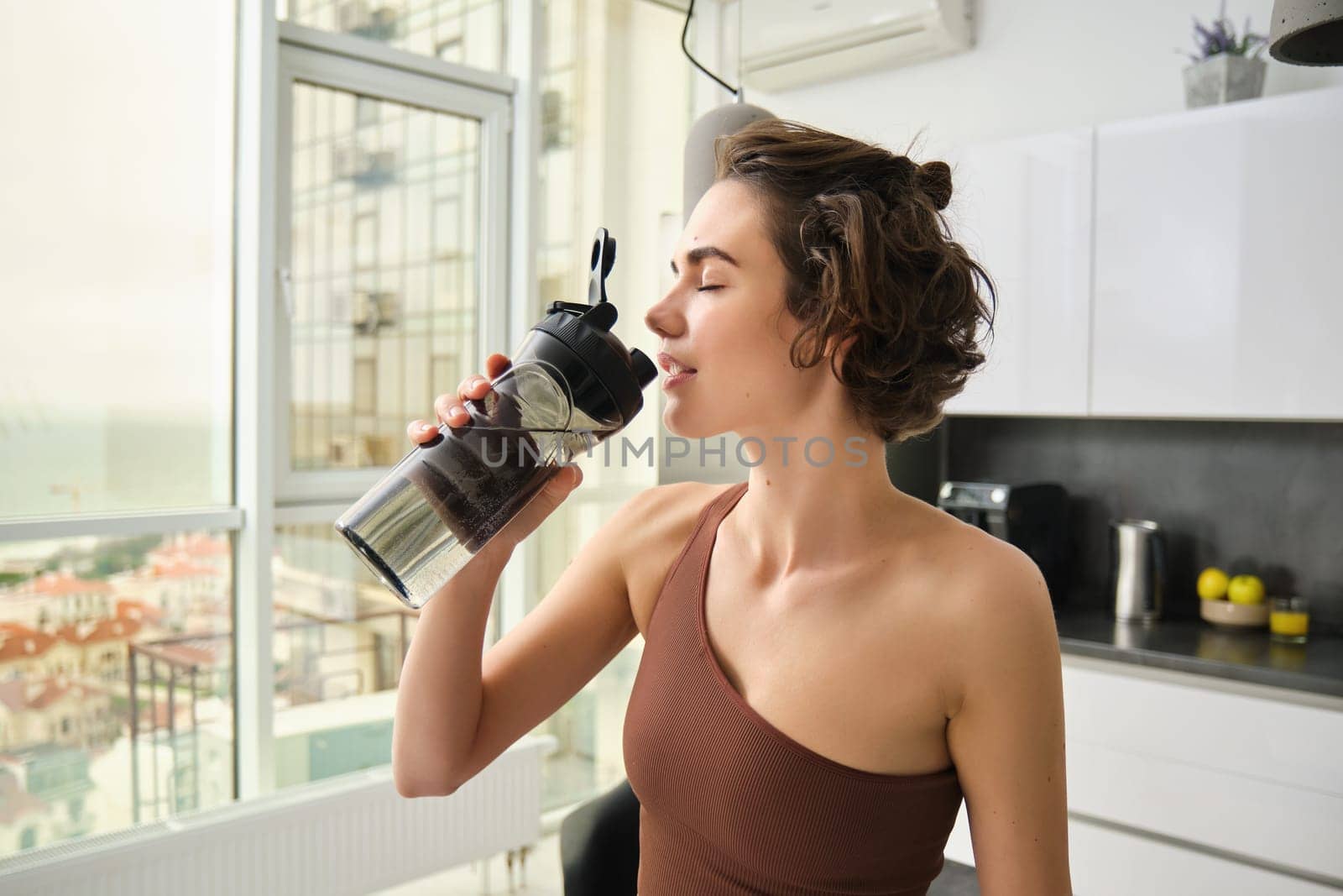 Female athlete in her kitchen, standing with water bottle, drinking after workout yoga training. Sportswear in sports wear stays hydrated after gym exercise by Benzoix