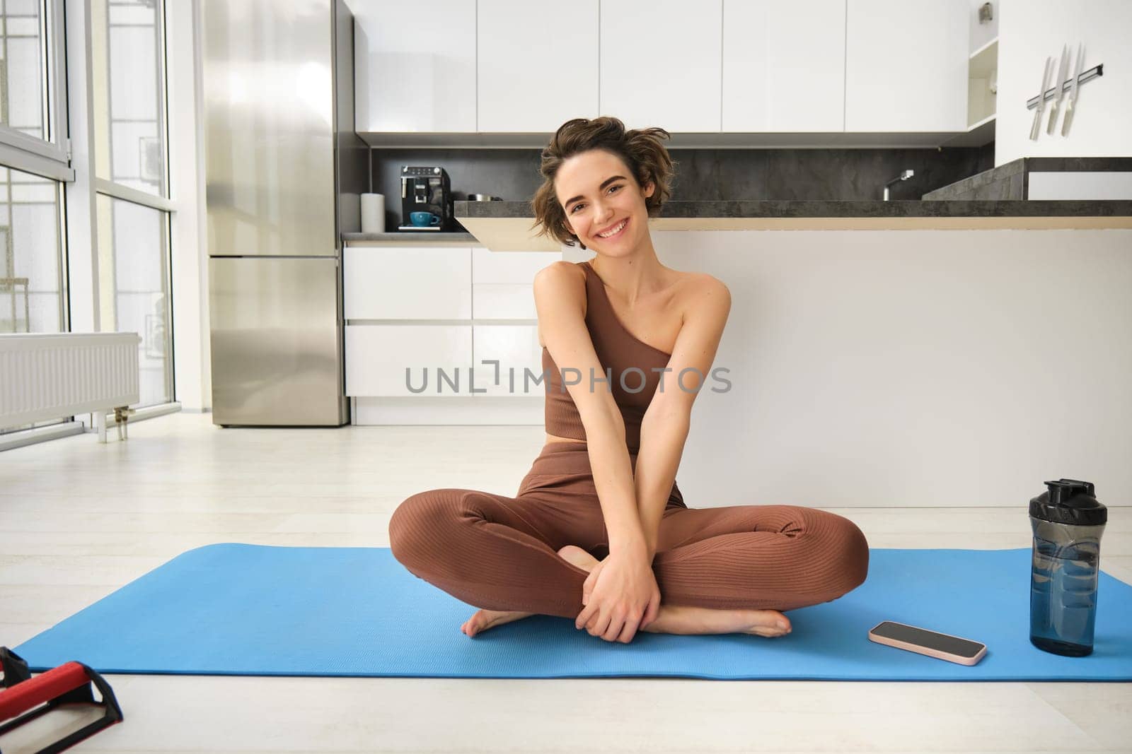 Portrait of fitness girl doing yoga on rubber mat at home, workout indoors in kitchen wearing sportswear, practice minfulness, sitting in lotus pose with water bottle next to her.