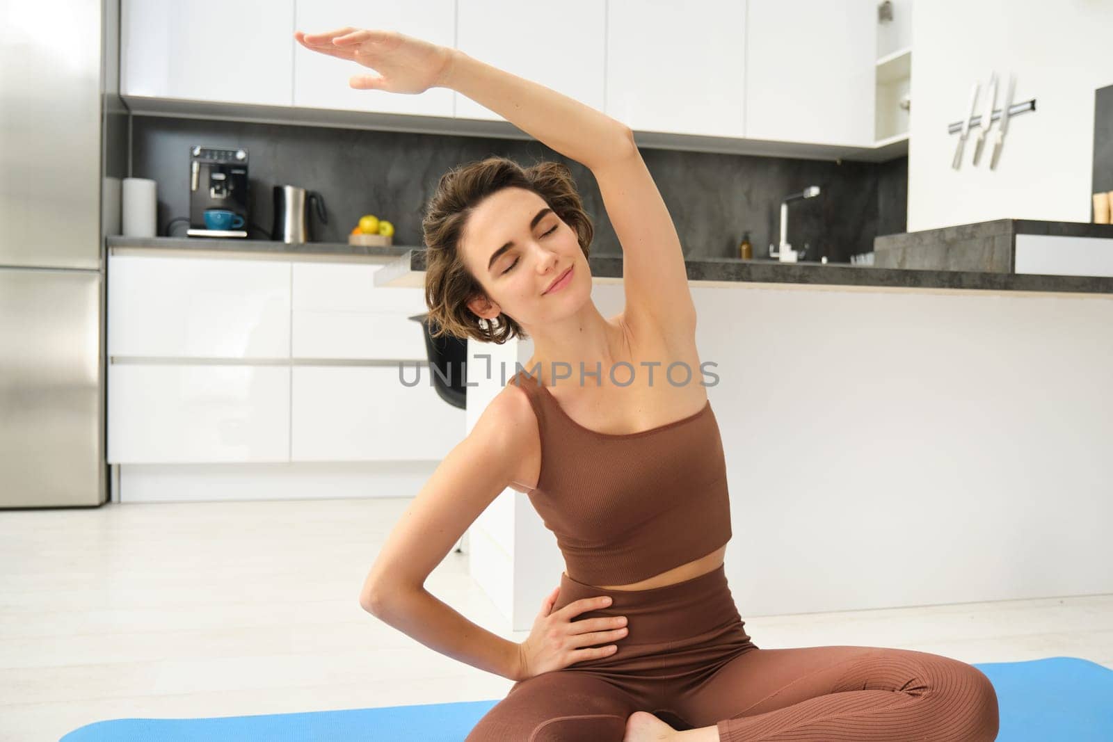 Portrait of fitness woman doing exercises in bright room at home, stretching in lotus pose, sitting on rubber yoga mat and doing workout training, drinking water from bottle by Benzoix