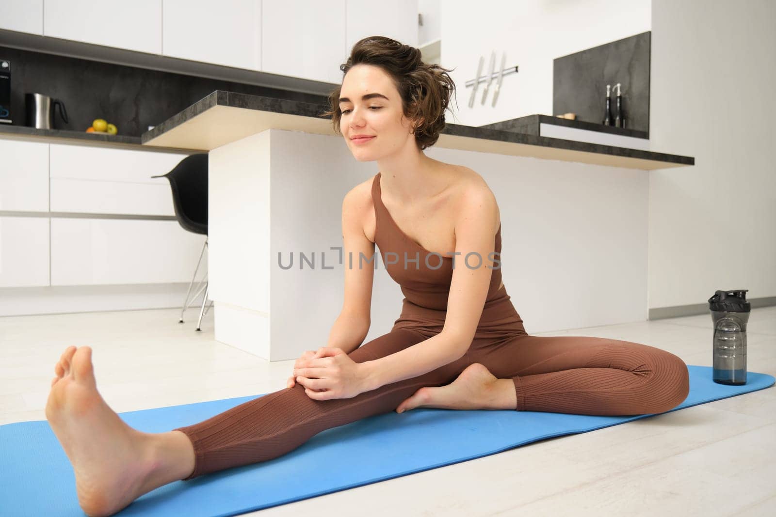 Portrait of brunette girl, fitness instructor, doing warm up training, exercises at home, stretching her legs on yoga mat at home by Benzoix