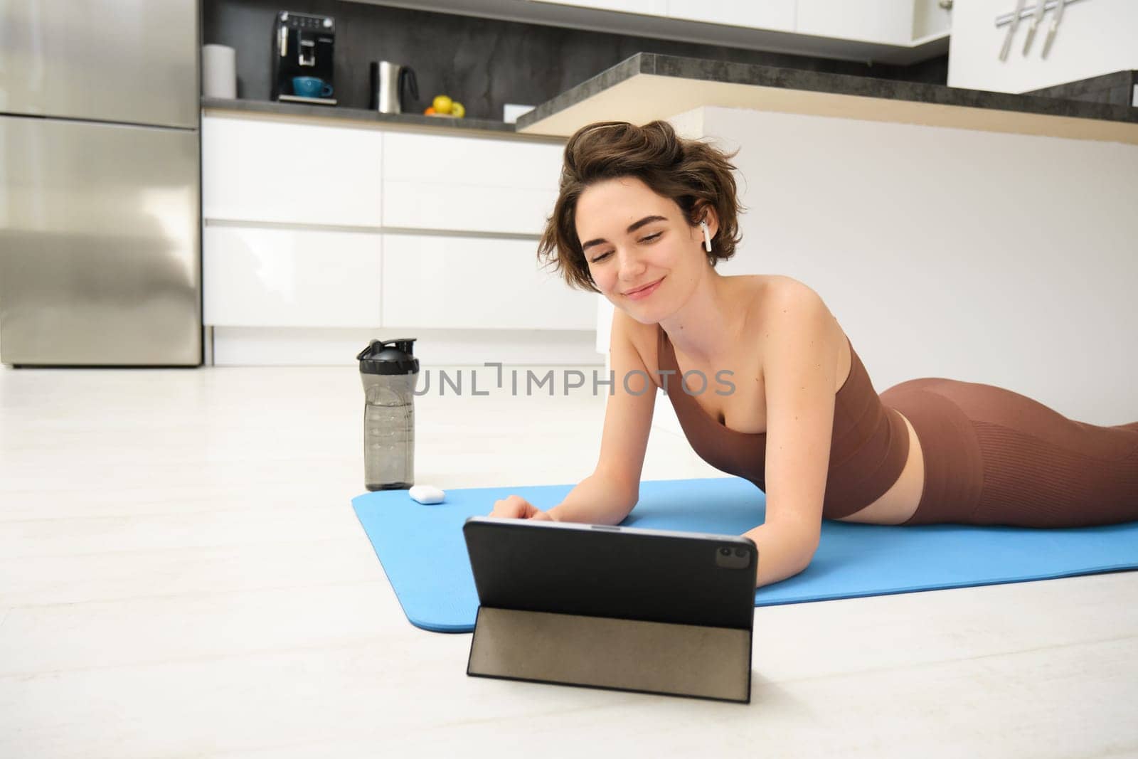Young fitness woman, girl in tracksuit lying on fitness mat, looking at yoga, pilates video tutorial on tablet, does workout from home, uses gym app, training at home by Benzoix