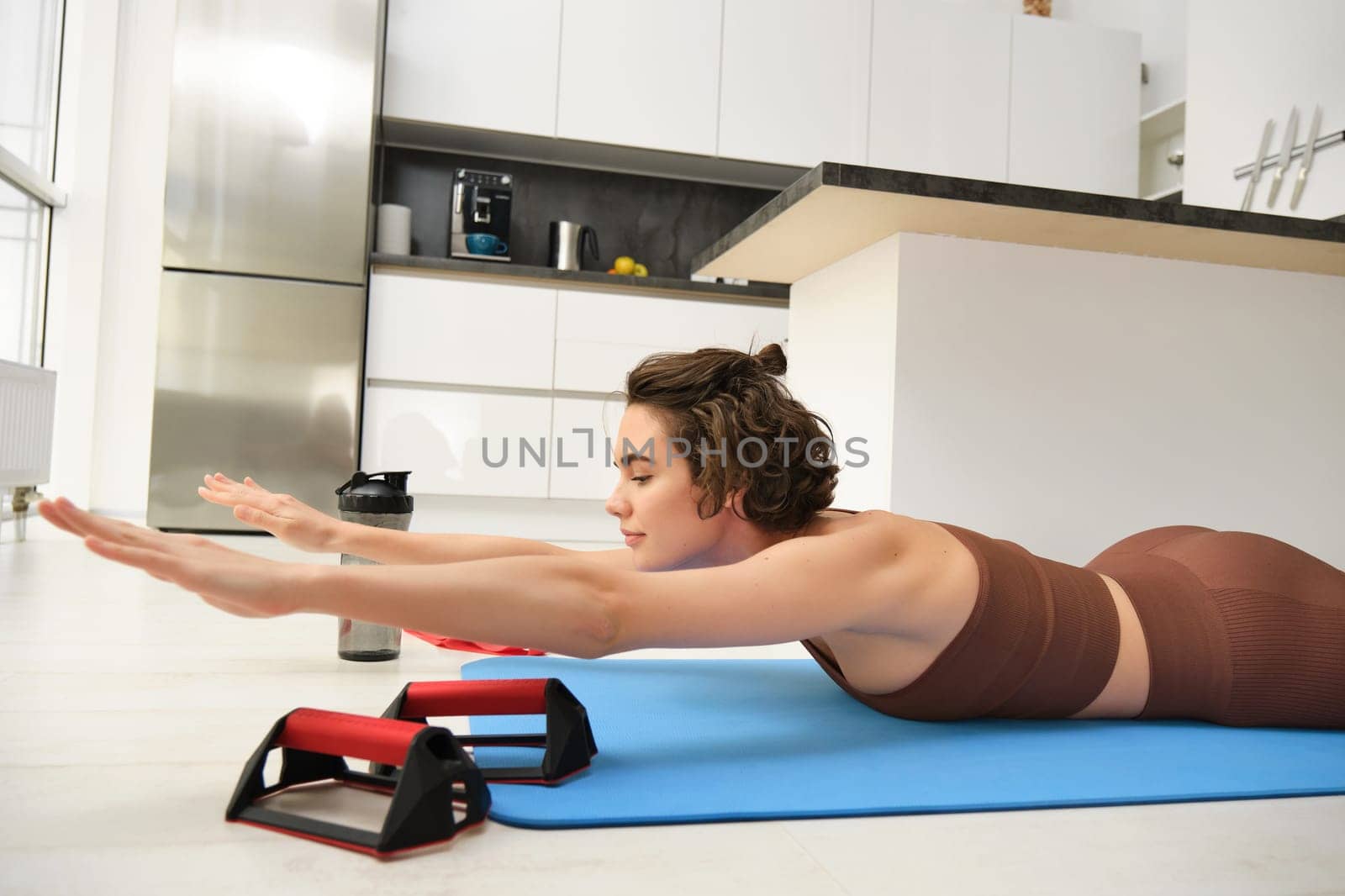 Portrait of young woman in activewear, stretching her arms, doing sports exercises at home, lying on rubber yoga mat and reaching hands forward, warm-up before workout.