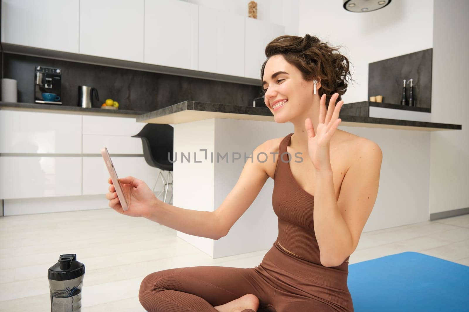 Young fitness girl at home, sport instructor in tracksuit waves hand at smartphone, sits on workout yoga mat, waves hand at mobile camera, says hi to online gym class, exercise remotely.