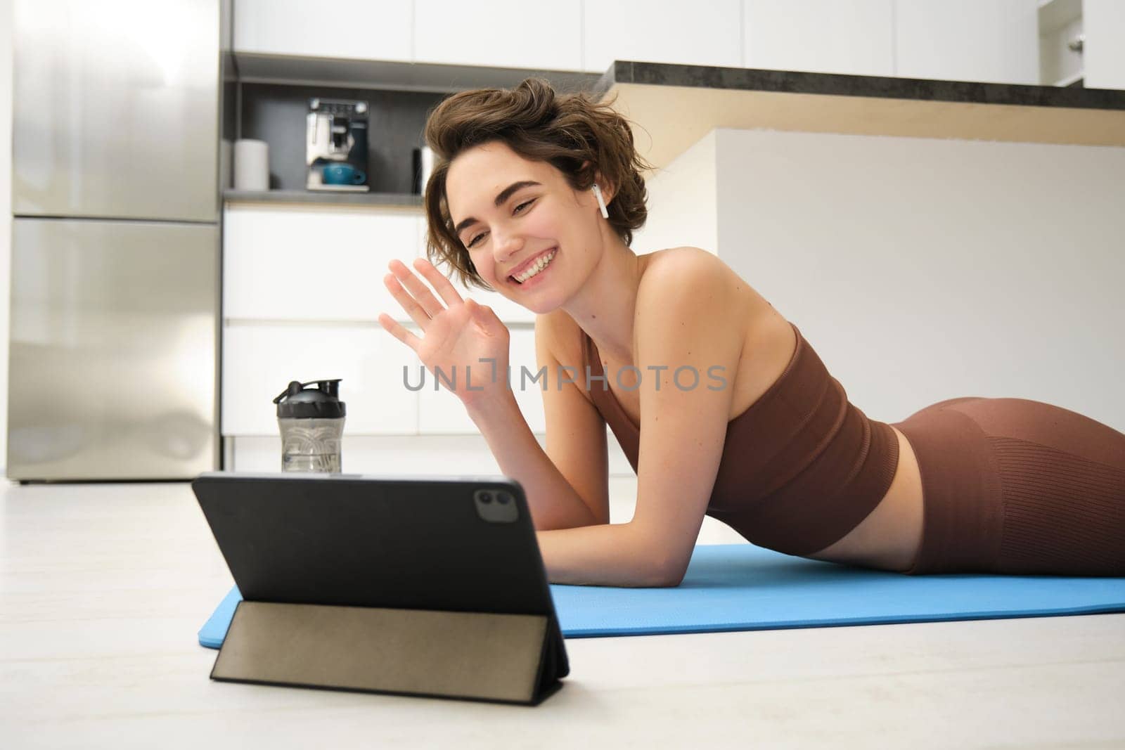 Smiling brunette woman in sportswear, waves hand, says hi at tablet, records video sports blog, fitness training session online, instructor shows workout elements, uses rubber mat at home in kitchen.
