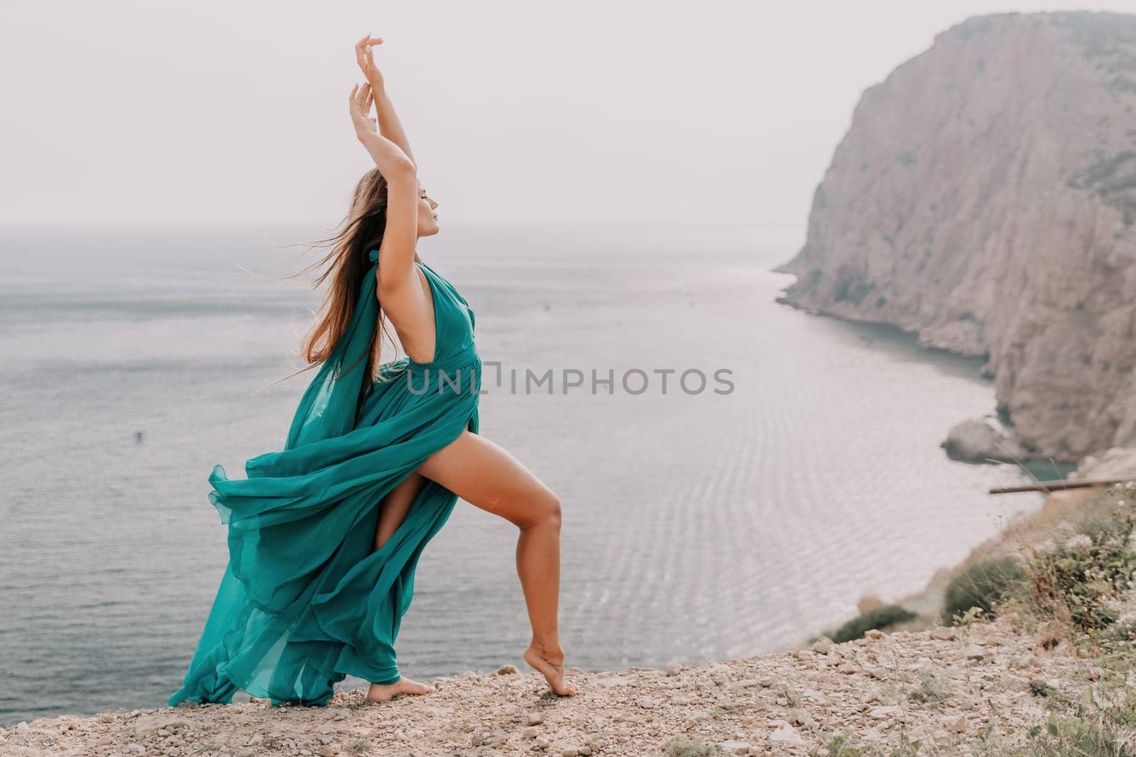 Woman travel portrait. Happy woman with long hair looking at camera and smiling. Close up portrait cute woman in a mint long dress posing on a volcanic rock high above the sea by panophotograph