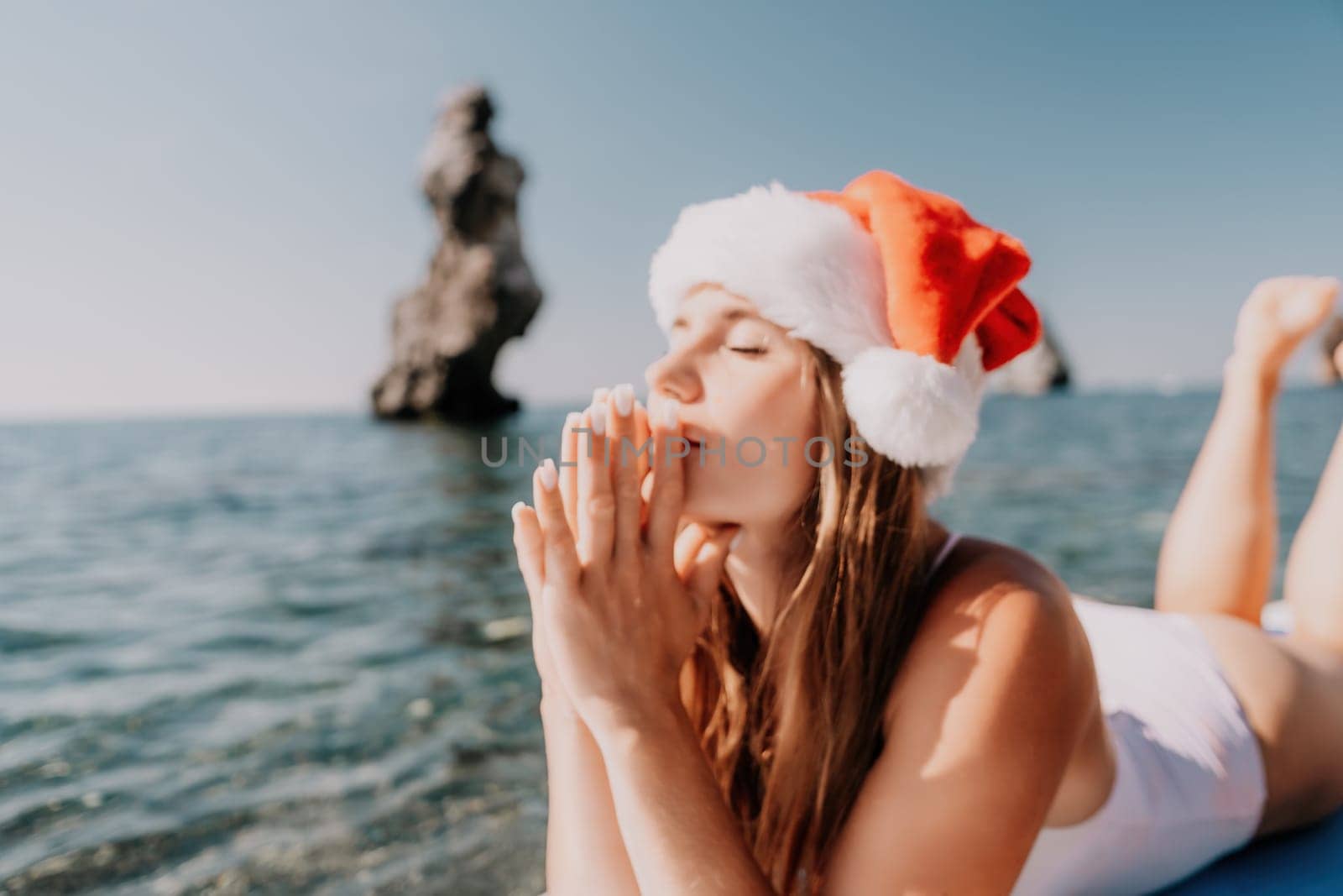 Woman sea sup. Close up portrait of happy young caucasian woman with long hair in Santa hat looking at camera and smiling. Cute woman portrait in a white bikini posing on sup board in the sea by panophotograph