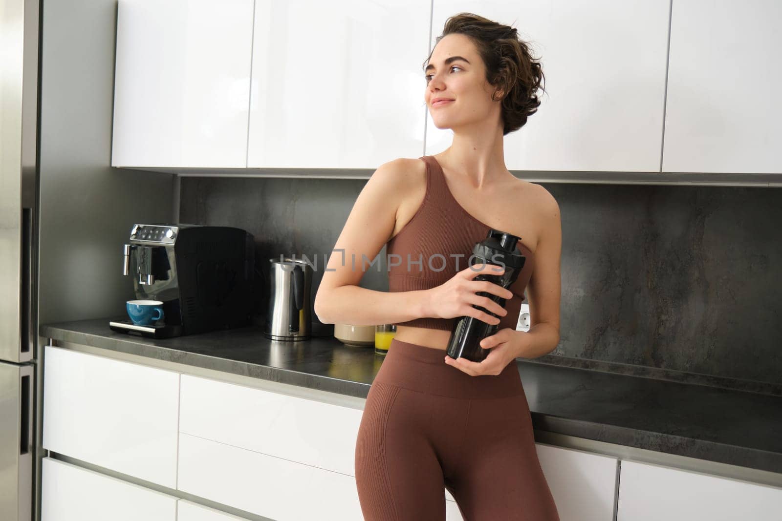 Portrait of smiling fitness girl, wearing sportswear for jogging, workout training, standing in kitchen and drinking water from sports bottle, looking fit and happy by Benzoix