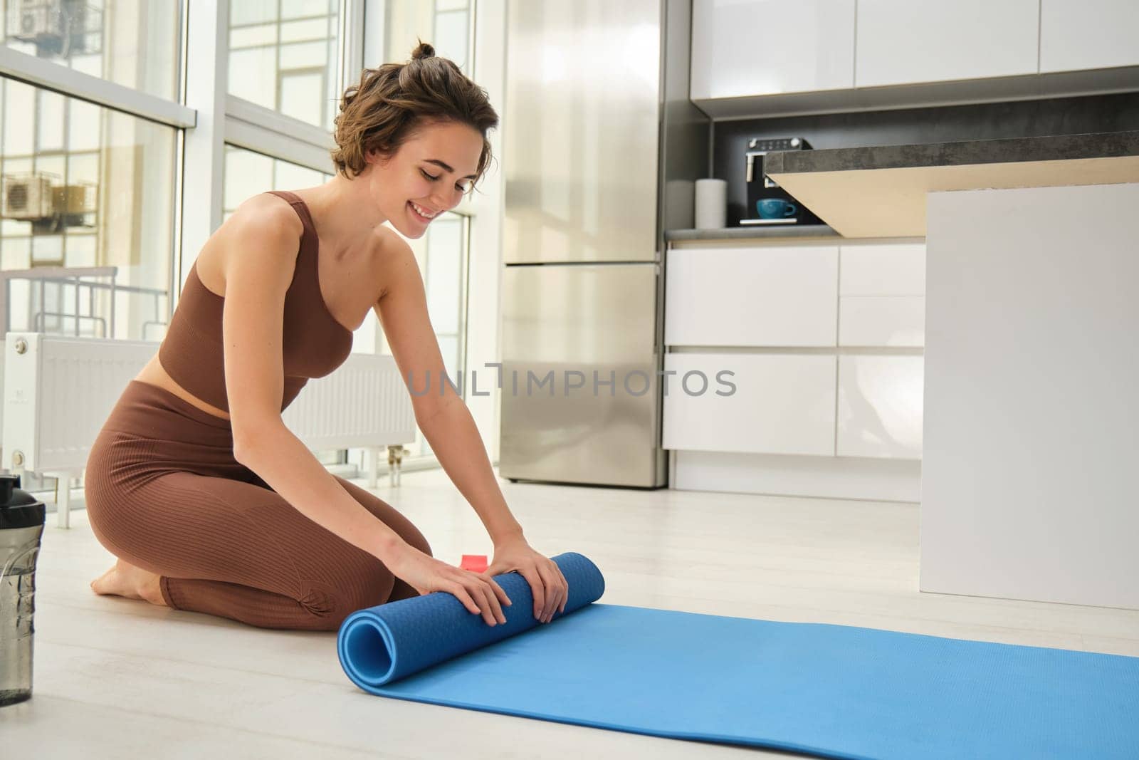Fitness and workout at home. Portrait of brunette sportswoman roll up her yoga rubber mat after finishing exercising in her kitchen. Young woman prepares space at her house for training pilates by Benzoix