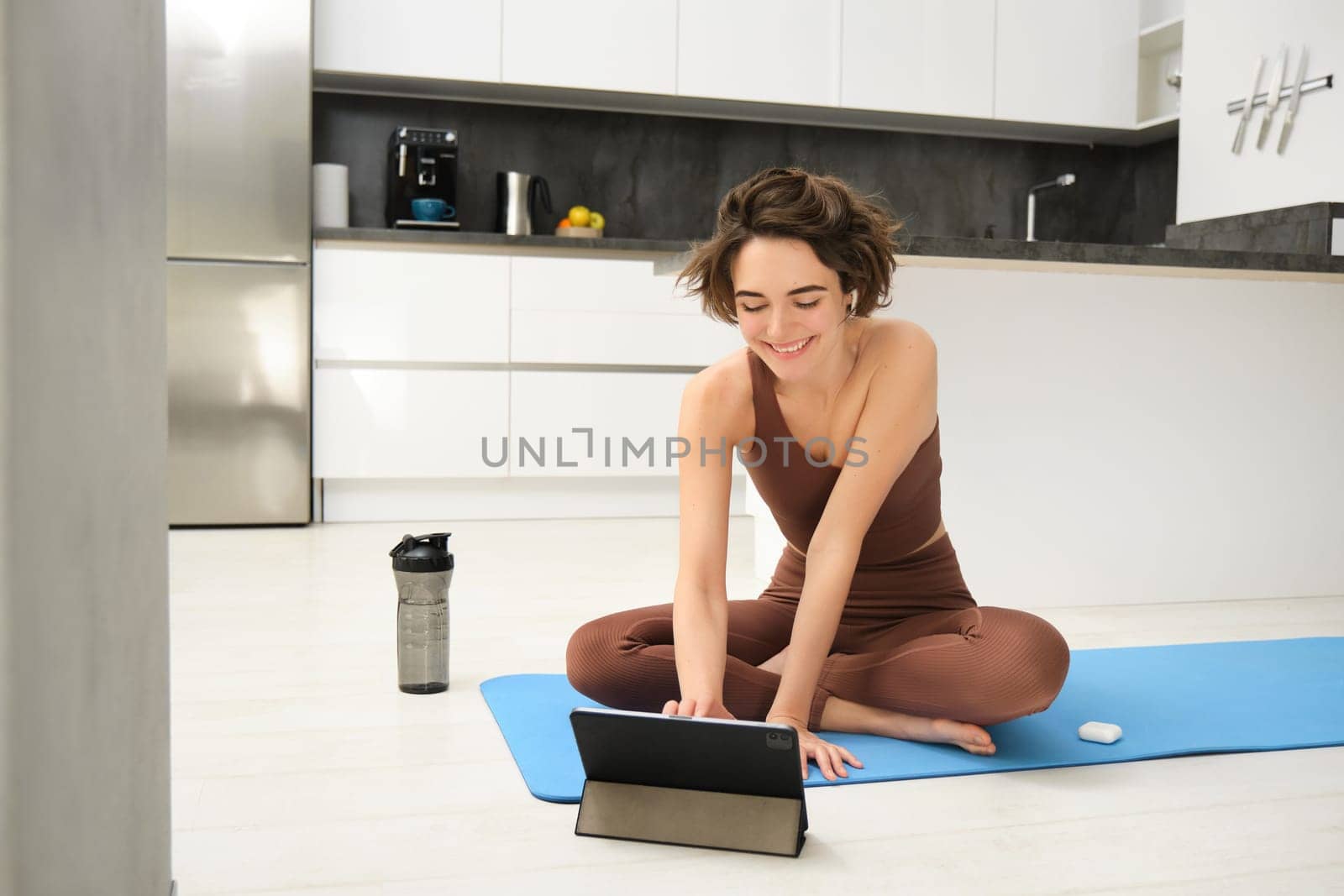 Young fit and healthy woman, yoga instructor, fitness girl in tracksuit, sits on floor rubber mat, using digital tablet, watches video pilates tutorial, stretching exercises by Benzoix