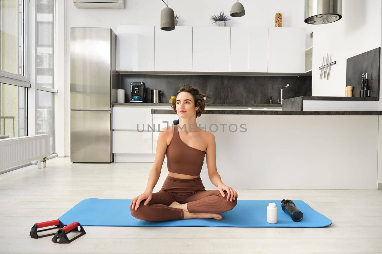 Portrait of young fitness woman, female athlete practice yoga from home, sitting in lotus pose with relaxed face, using rubber mat in kitchen for workout by Benzoix