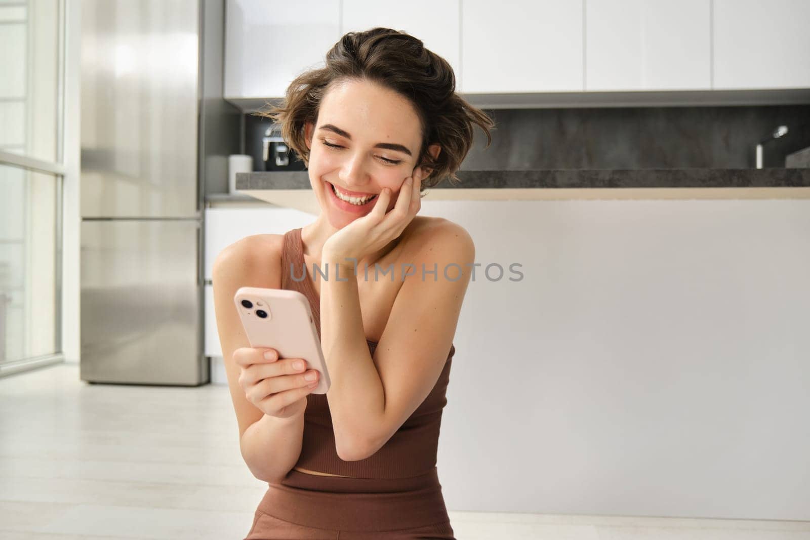Portrait of smiling woman at home, using her mobile phone, sitting on floor in bright room, looking at smartphone and chatting, texting someone. Copy space