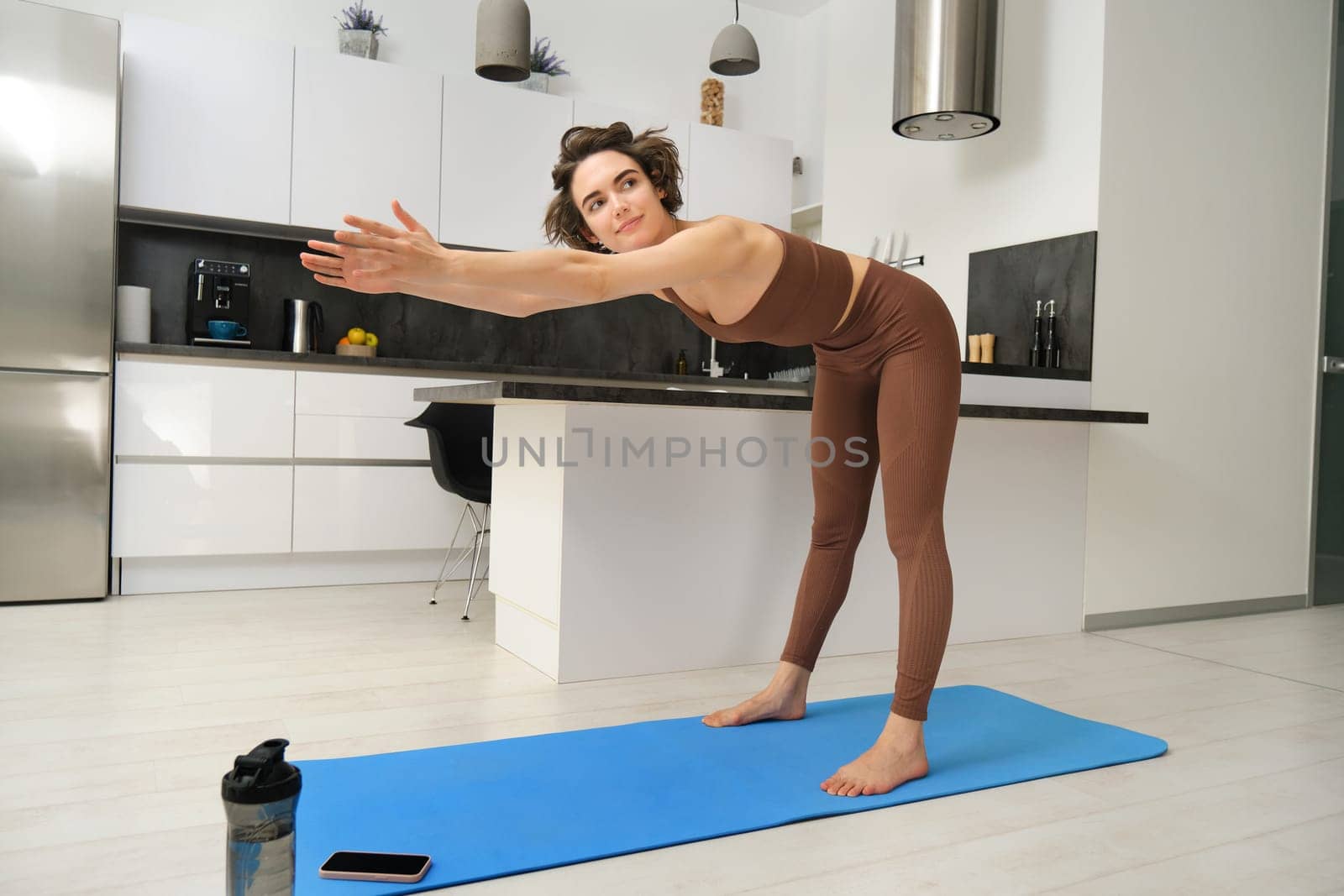 Woman follows gym video tutorial on smartphone while workout at home. Fitness girl does training at home on rubber yoga mat by Benzoix