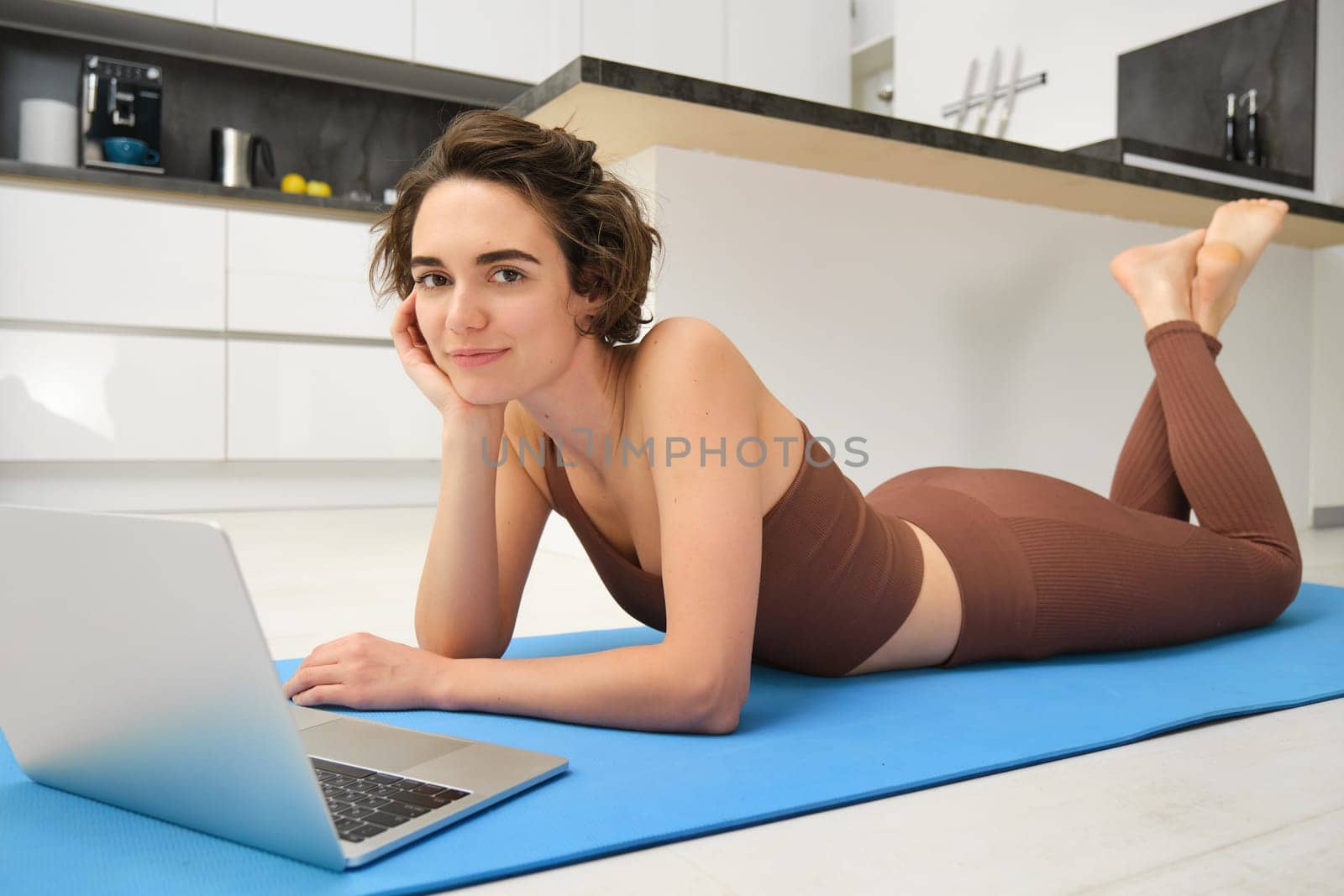 Portrait of beautiful sportswoman, fitness girl watching videos on laptop during workout, follow online pilates videos while exercise at home, wearing activewear in kitchen.