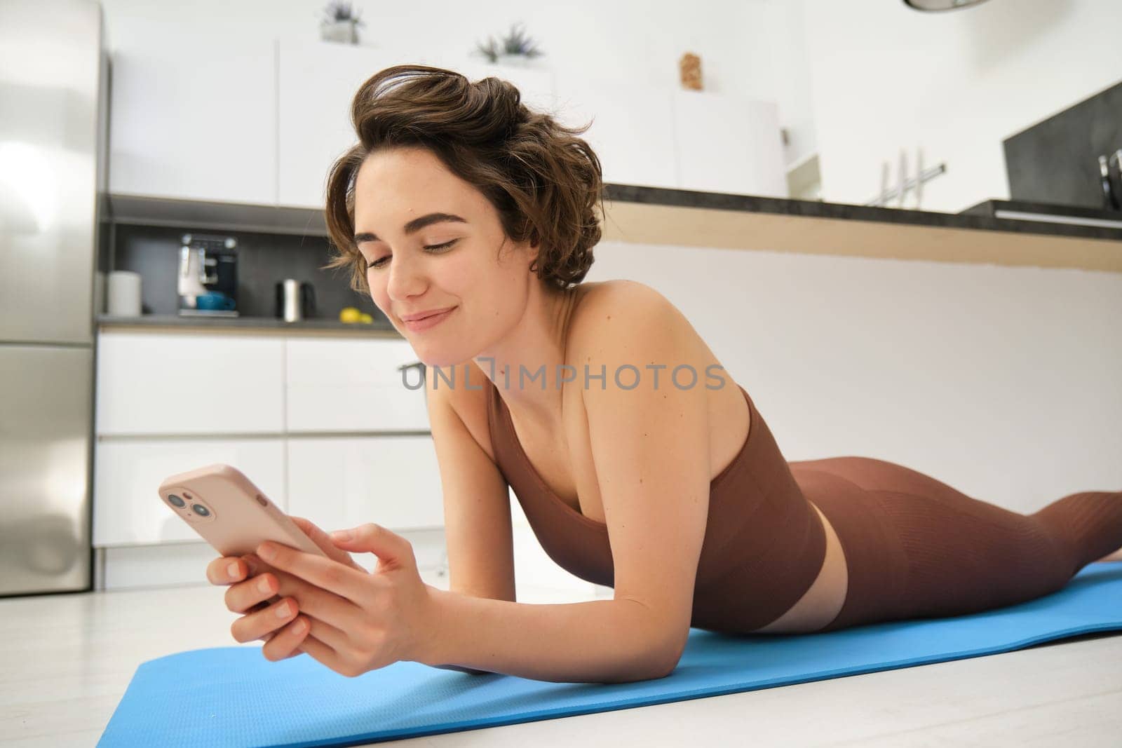 Image of sporty girl resting on yoga rubber mat, workout at home, using her smartphone, looking for online fitness tutorials, mobile app for training.
