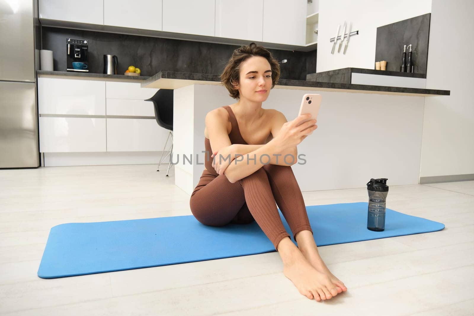 Portrait of fitness girl in sportswear, looking at mobile phone, workout from home, looking for exercise instructions online on smartphone app by Benzoix