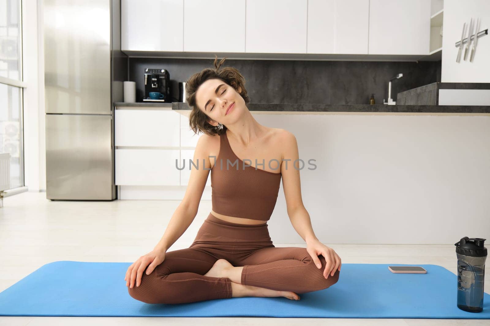 Photo of focused young woman in sportswear stretching her neck while working out on mat at home, doing fitness exercises.