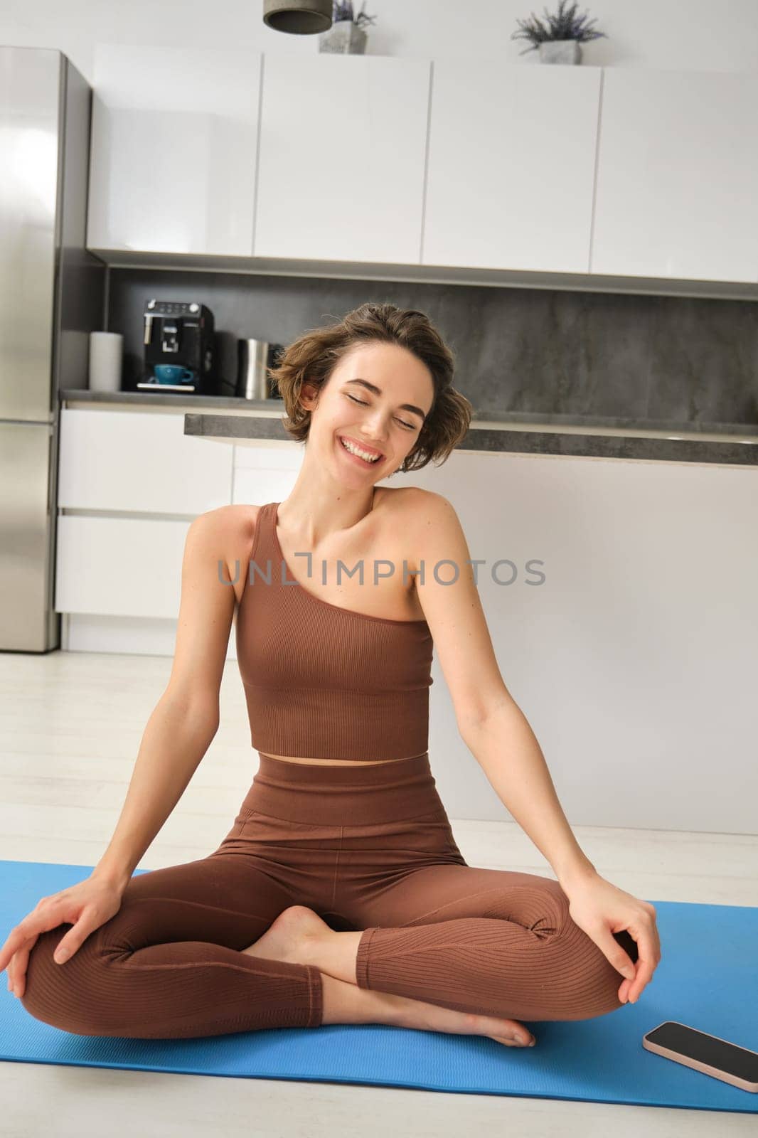 Vertical shot of smiling brunette fitness woman, young athlete meditating, doing yoga at home on floor, practice mindfulness by Benzoix
