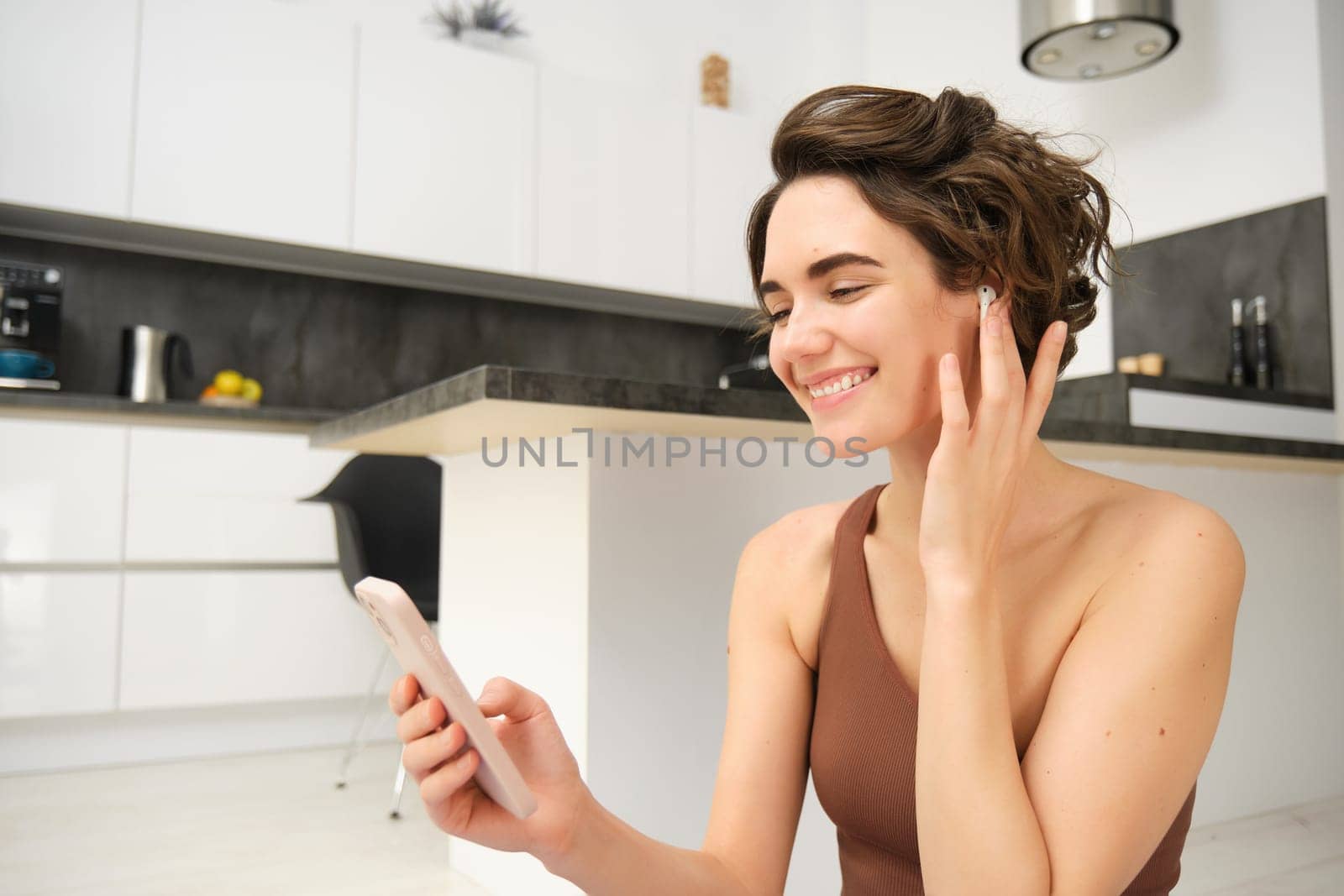 Close up of young fitness instructor, healthy and fit woman with smartphone, wireless headphones, records her workout training session at home, connects to client on video call for yoga classes by Benzoix