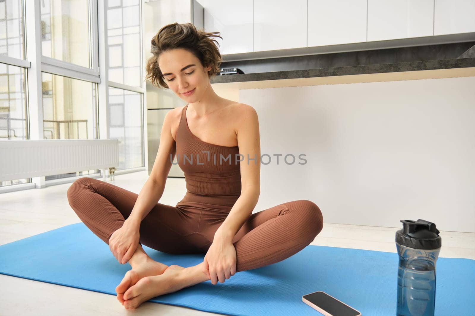 Sport and wellbeing. Young brunette fitness girl, wearing activewear, sitting on yoga mat, meditating, practice relaxation and mindfulness at home, workout in kitchen by Benzoix