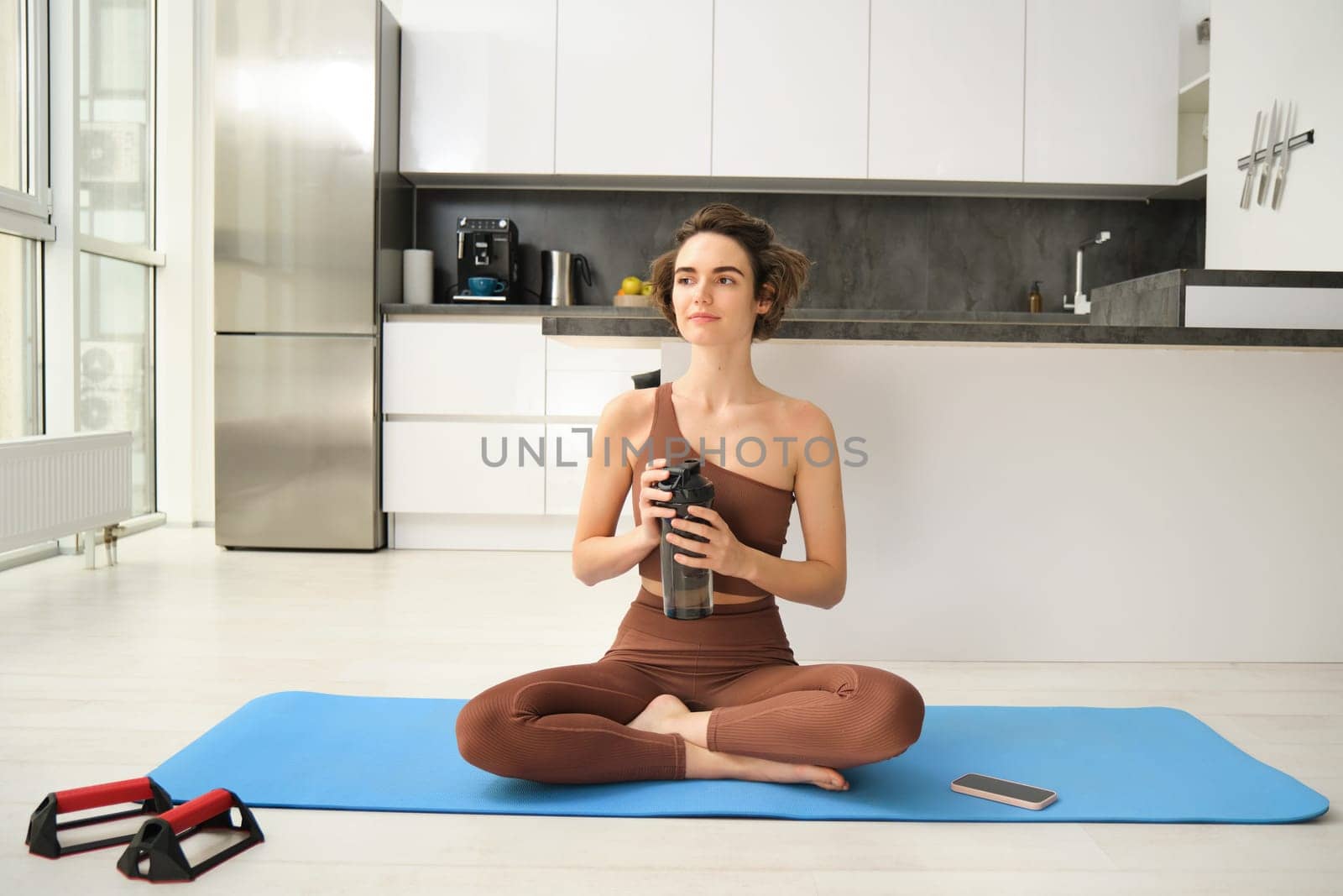Young strong sporty athletic fitness trainer, instructor woman wearing brown tracksuit sitting on yoga mat drink water training do exercises at home gym indoor. Workout sport motivation concept.