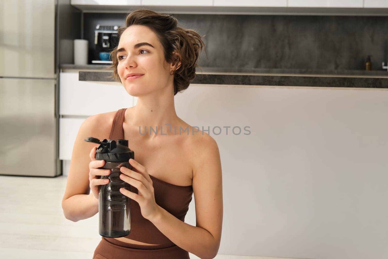 Fitness instructor, smiling young woman workout at home, sits on yoga mat and drinks water, does exercises, training session in her kitchen by Benzoix