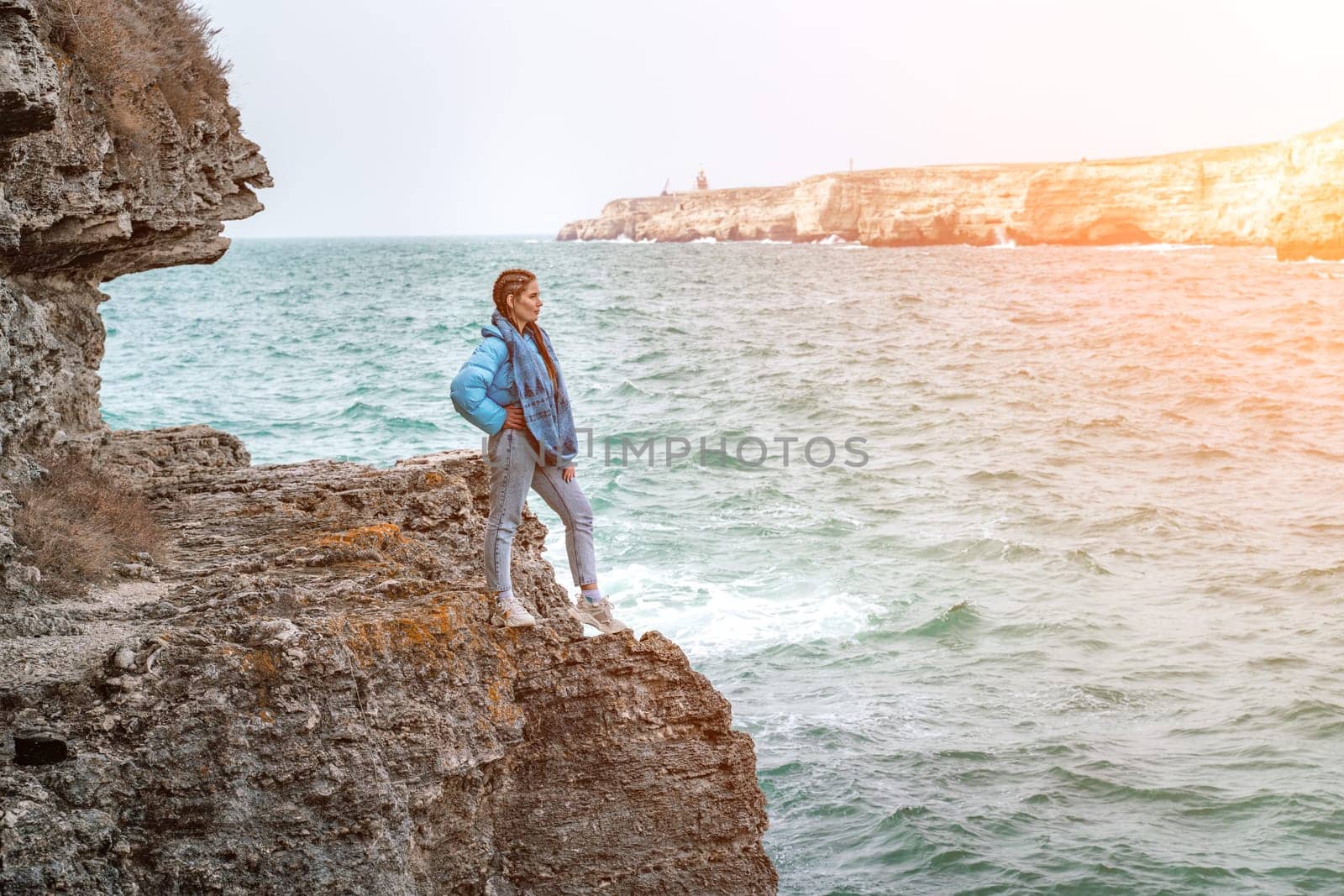 woman sea travel. A woman in a blue jacket stands on a rock above a cliff above the sea, looking at the stormy ocean. Girl traveler rests, thinks, dreams, enjoys nature.