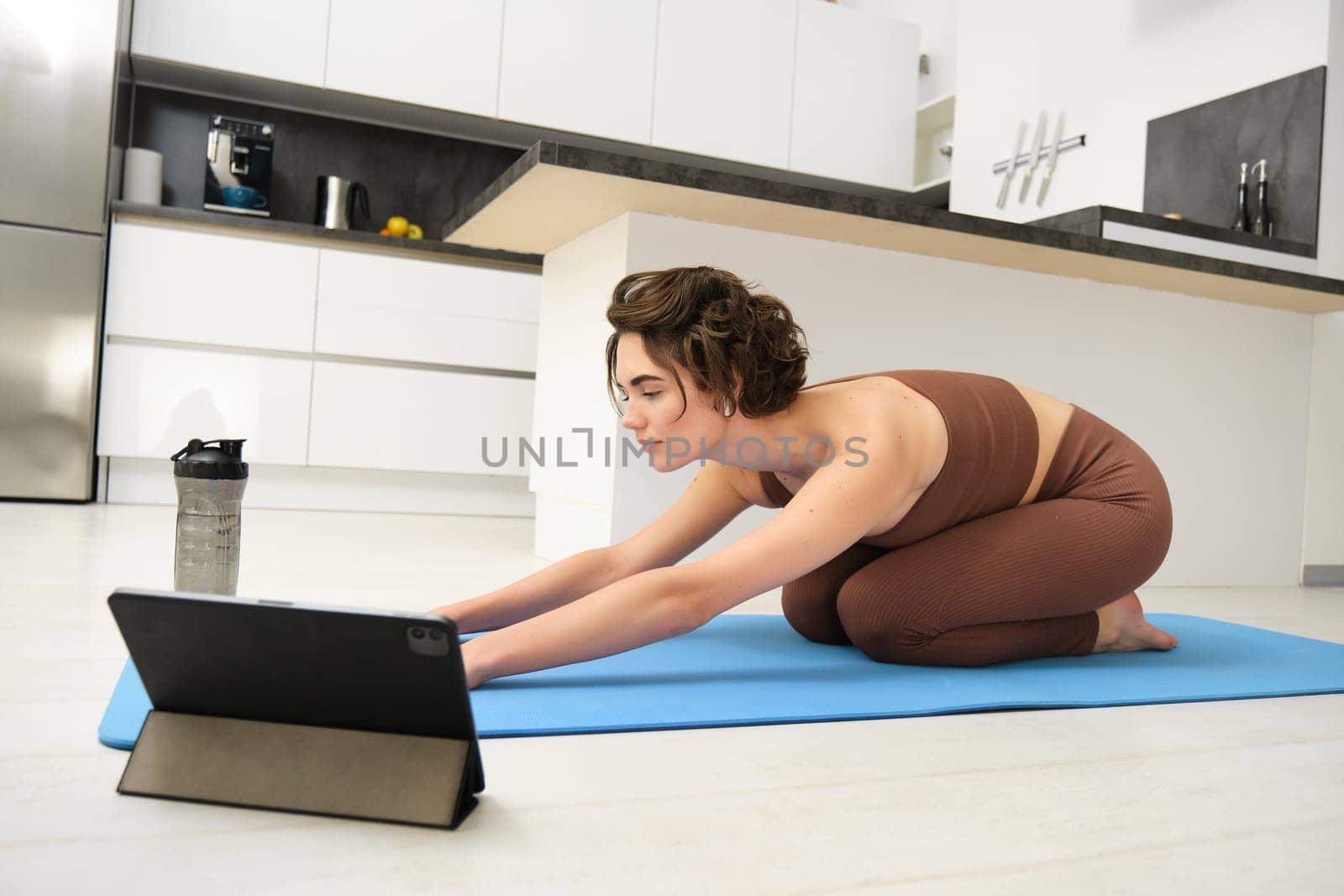 Image of young sportswoman does yoga at home, watches gym instructor tutorial video on tablet, stretches body, fitness training exercises, pilates class online from home by Benzoix