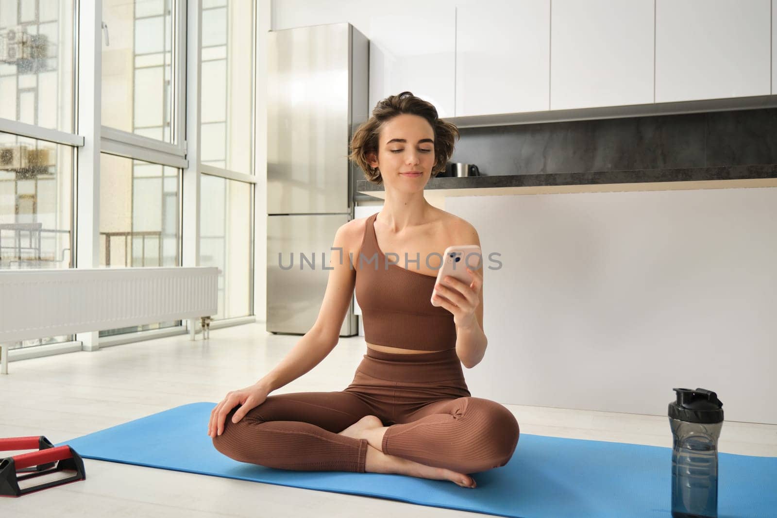 Sport app and wellbeing. Young woman, athlete sitting on floor at home with smartphone, doing yoga, using workout app on mobile phone, doing exercises at home by Benzoix