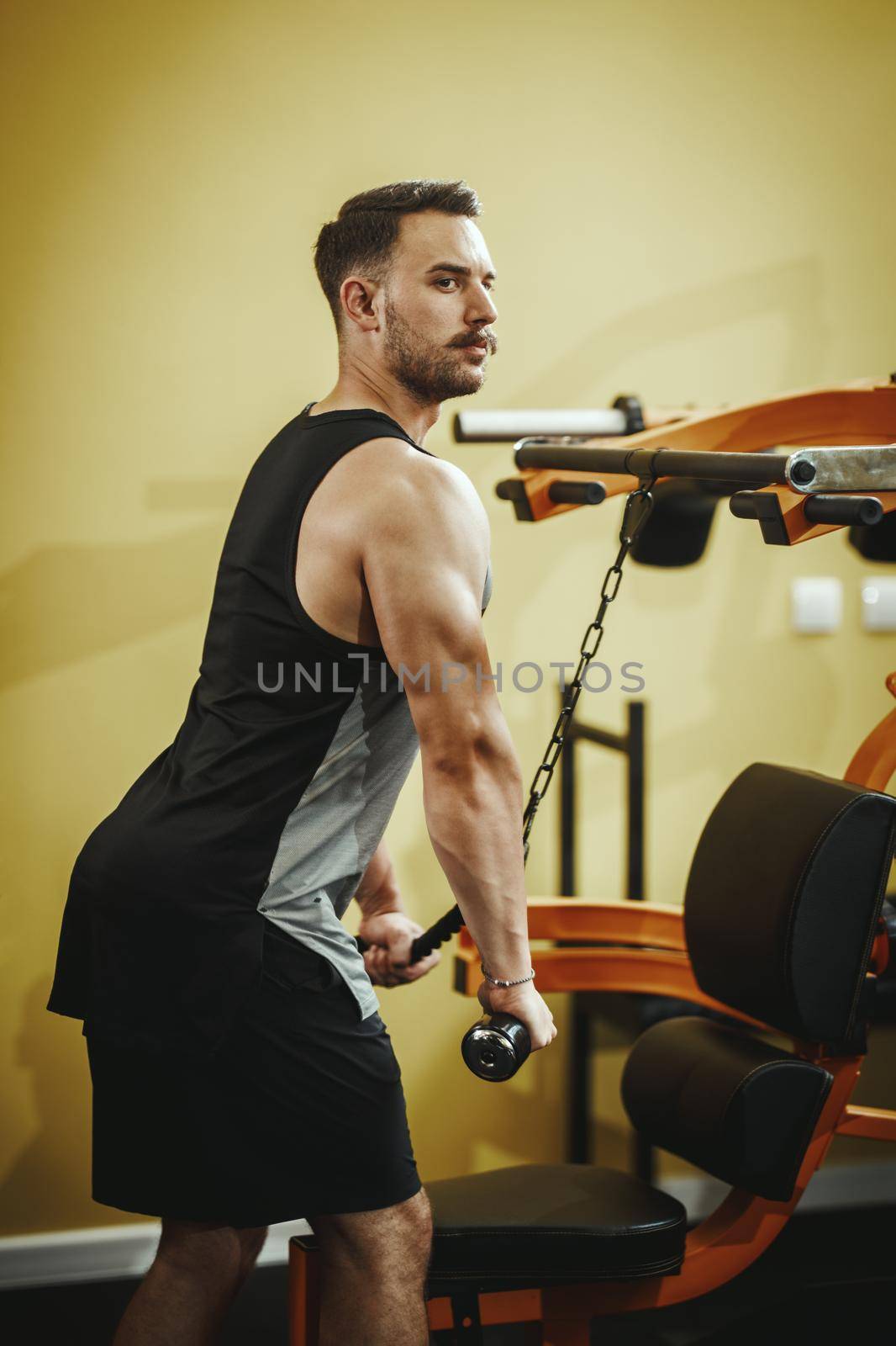 Shot of a muscular guy in sportswear working out on machine at the cross training gym. He is pumping up triceps muscule with heavy weight.