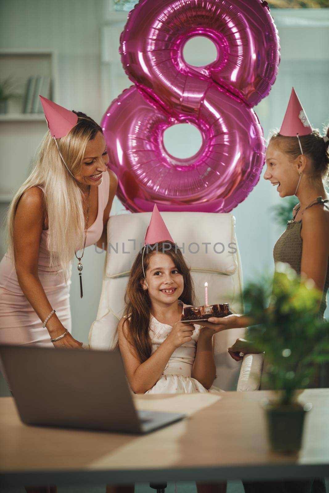 Beautiful young mom with her daugters at home during pandemic isolation have birthday party, she blowing candles and have conference call with friends and family.