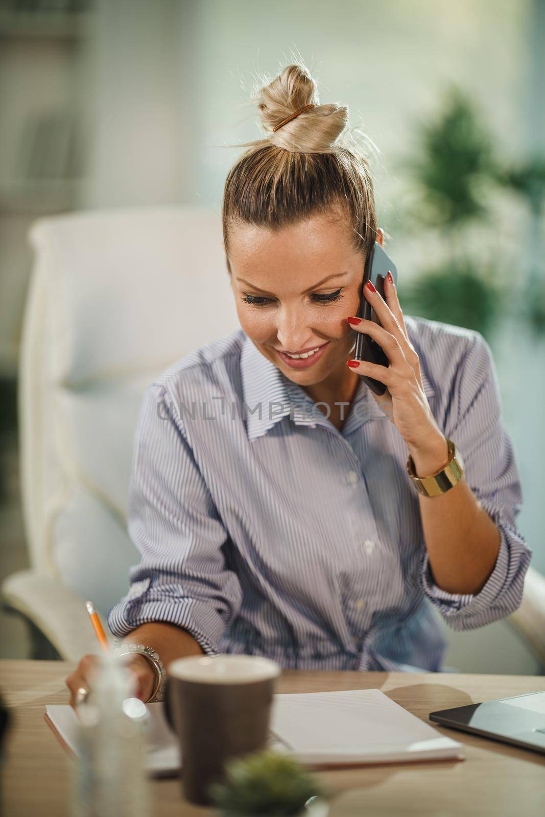 Shot of a attractive young businesswoman sitting alone in her home office and talking on smartphone during COVID-19 pandemic.