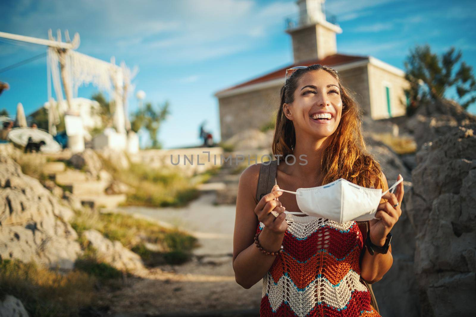 Shot of a smiling young woman putting on a protective mask and spending time on seaside, exploring a Mediterranean at corona pandemic. 