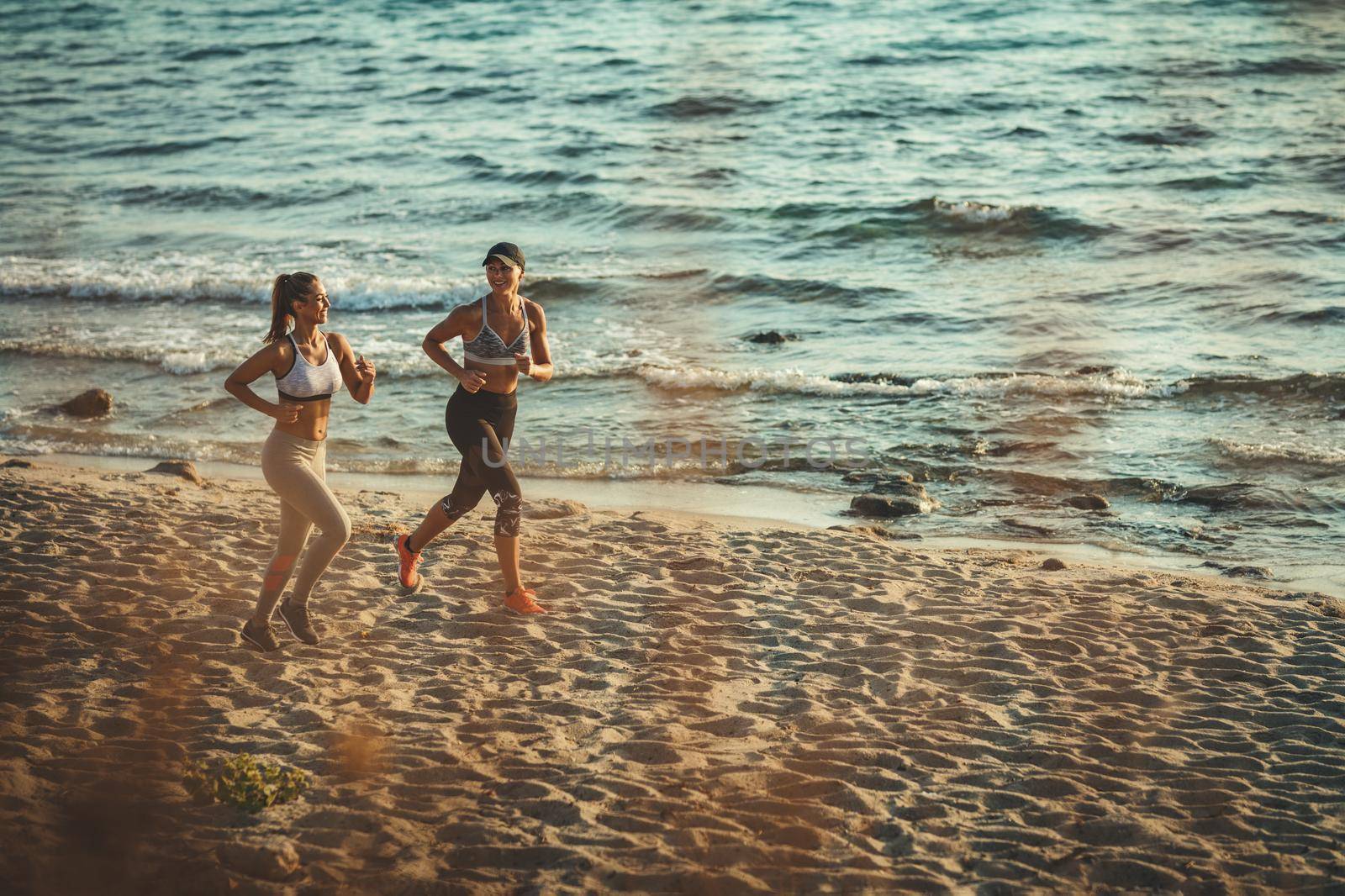 Two smiling happy young women are running along the sea shore on the beach during the hot summer vacation day.