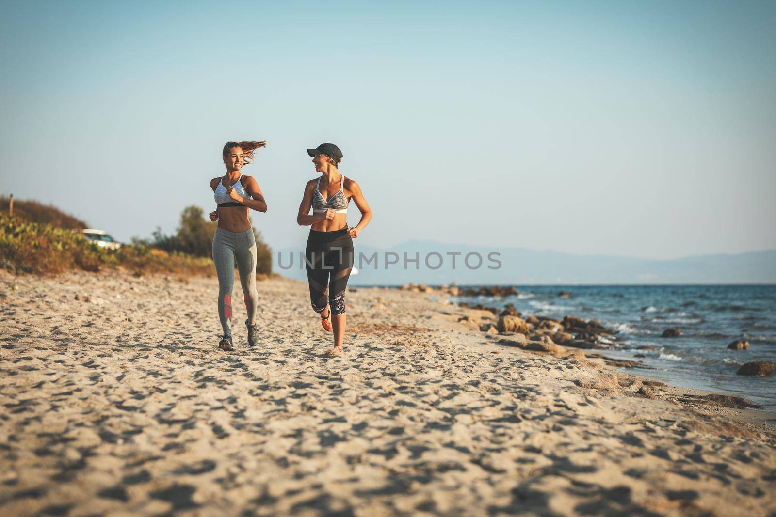 Two smiling happy young women are running along the sea shore on the beach during the hot summer vacation day.