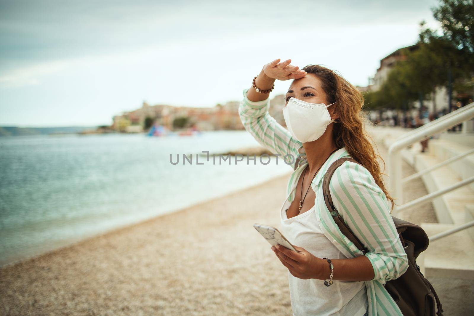 Shot of an attractive happy young woman wearing protective N95 mask and using smartphone while enjoying a vacation on the beach during the COVID-19.