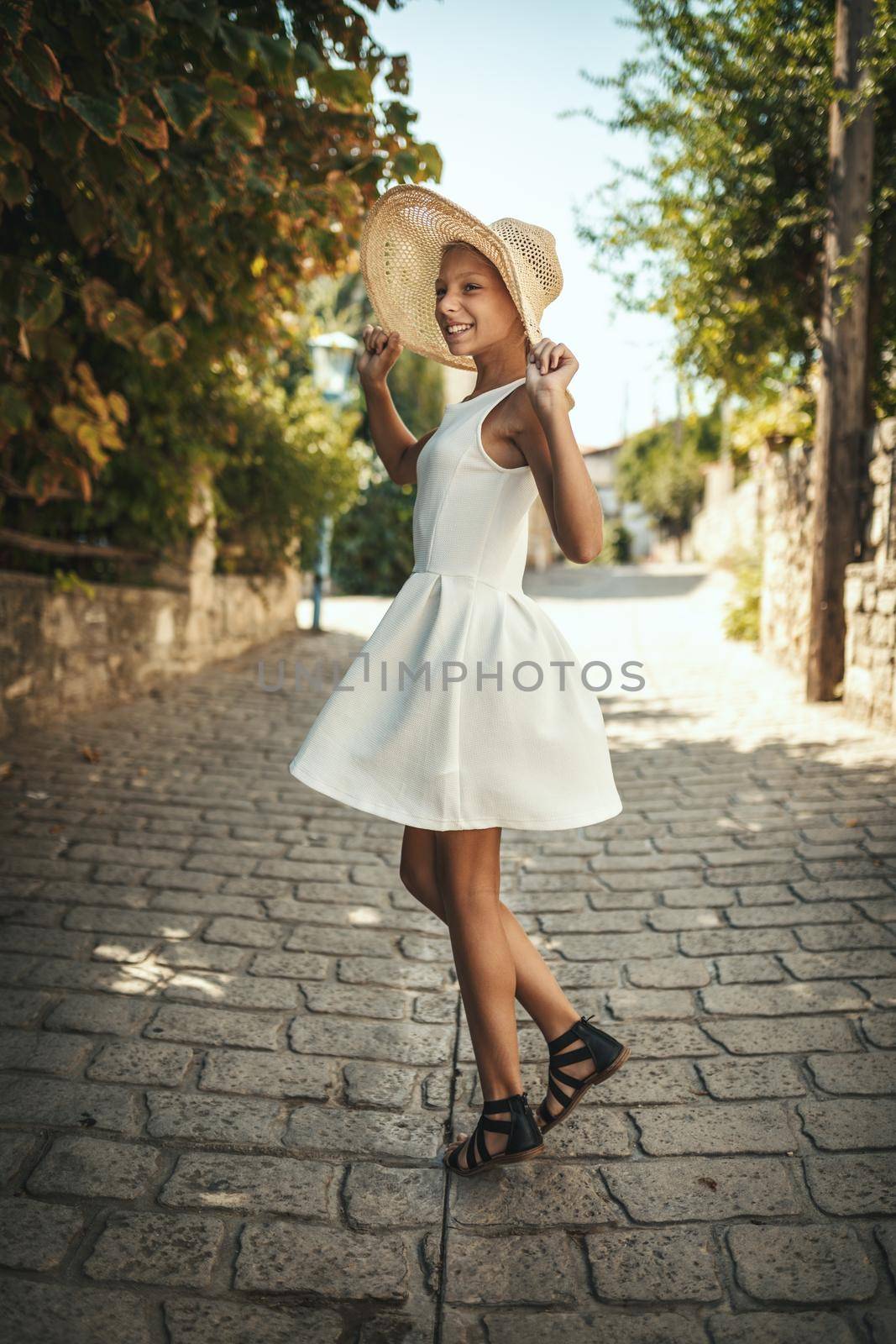 A beautiful teenage girl is walking along the streets of a Mediterranean town and enjoys in summer sunny day. She is smiling, having fun and dancing with straw hat on her head.