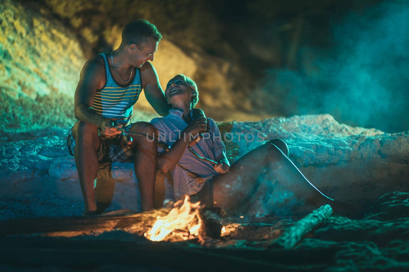 The happy smiling couple is sitting on the sandy seashore by the fire in the evening. They are frying sausages on stick.