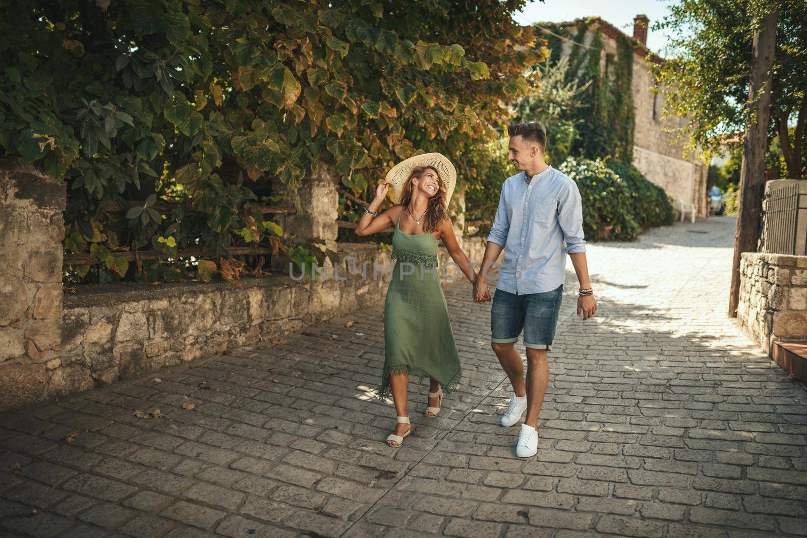 A beautiful young couple is having fun and dancing on the street while walking along of a Mediterranean town. They are enjoyed in summer sunny day, holding each other's hand and smiling.