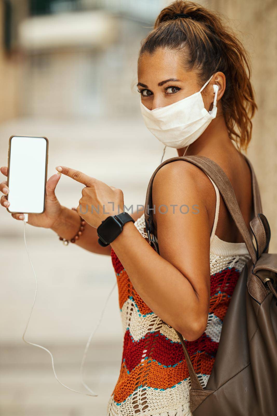 Shot of a happy young woman wearing protective mask and pointing on her empty smartphone screen during spending time on vacation and exploring a Mediterranean city at corona pandemic. 
