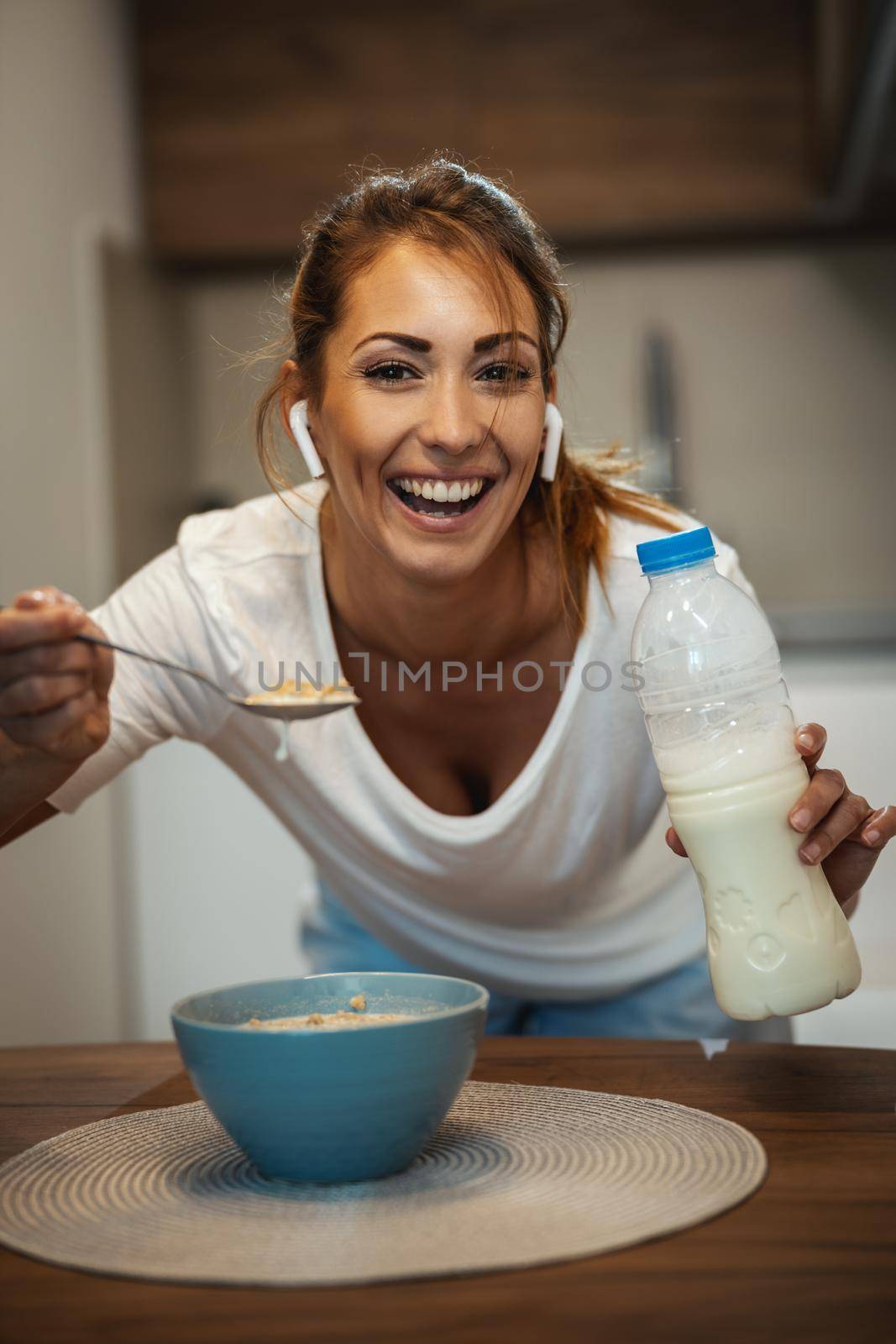 Beautiful young smiling woman is eating her healthy breakfast in her kitchen and listening music. Looking at camera.