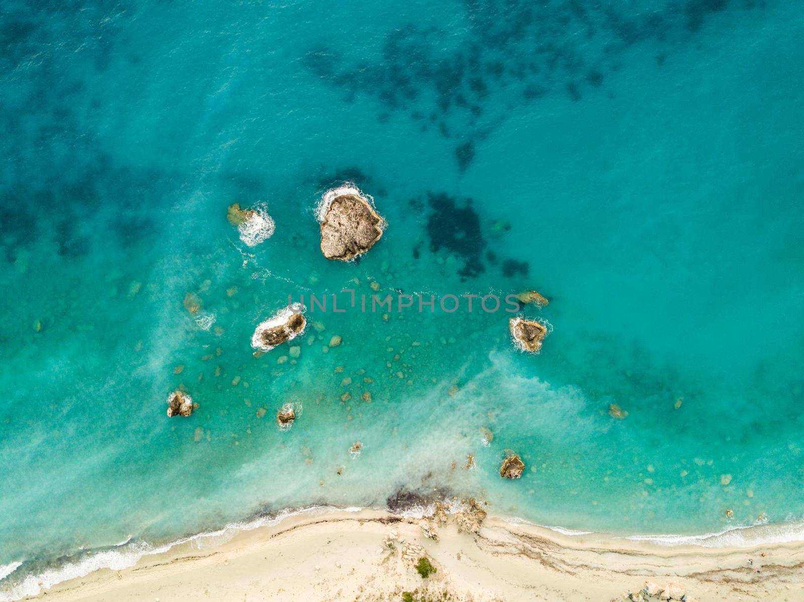 Aerial view from drone of the beautiful crystal clear turquoise and blue sea water, surrounding rocks and waves reaching sandy beach. 