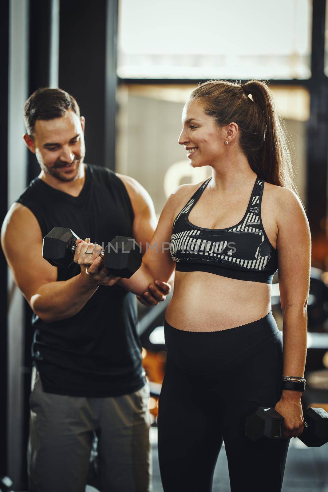 Shot of a pregnant woman working out with a dumbbell under control of the personal trainer at the gym.