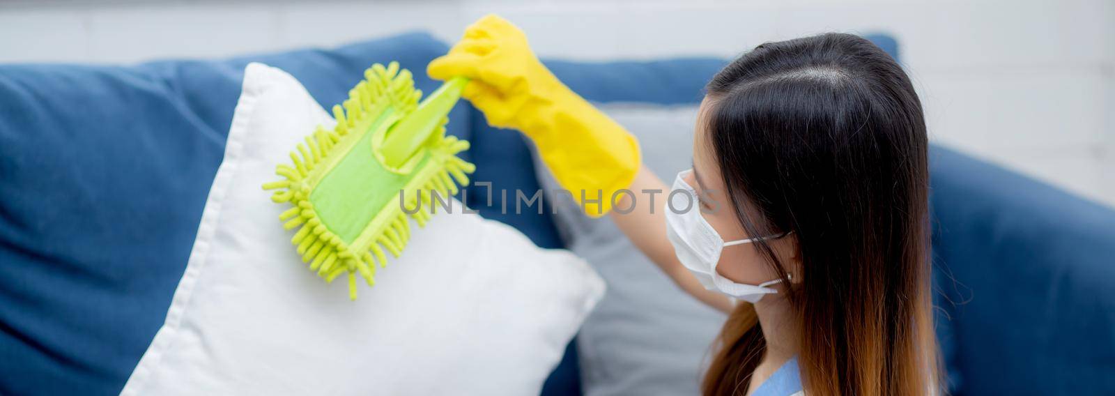 Young asian woman in face mask and gloves cleaning dust with duster on sofa and cushions at home, girl doing housework for hygiene at house, housekeeper and service, health care and lifestyle concept. by nnudoo