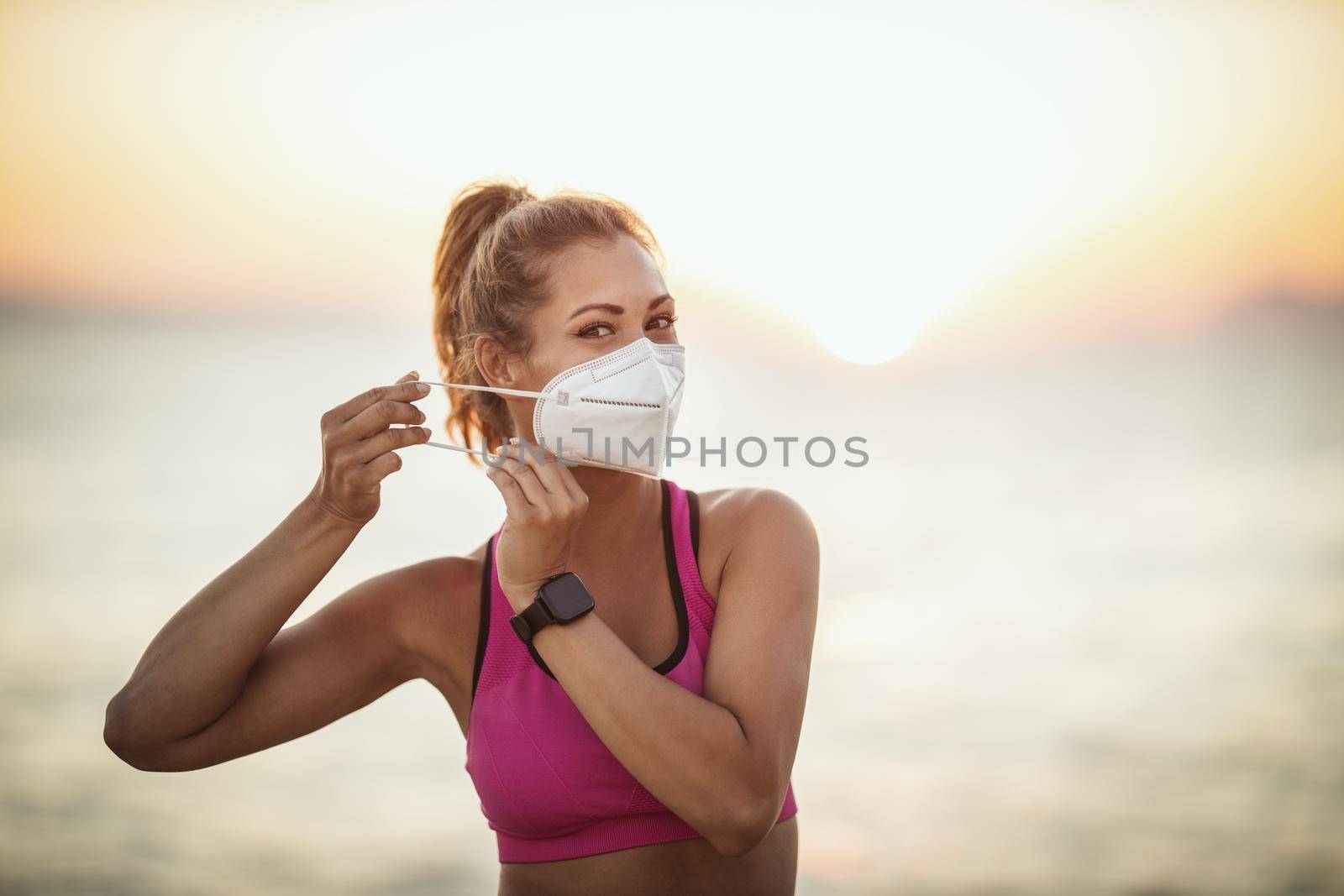 Portrait of a female runner wearing protective face mask while doing training near the beach at sunset during the COVID-19 pandemic.