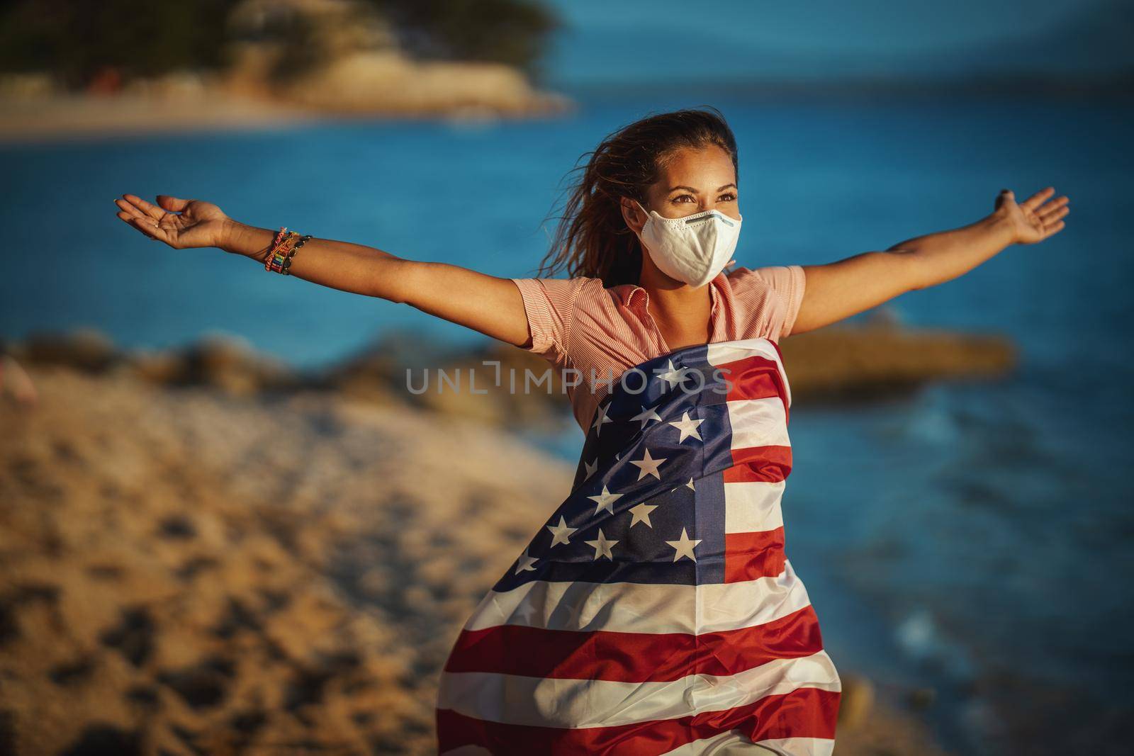 Attractive happy young woman with US national flag and protective N95 mask on the beach during preventing the spread of the epidemic and treating coronavirus and pandemic COVID-19.